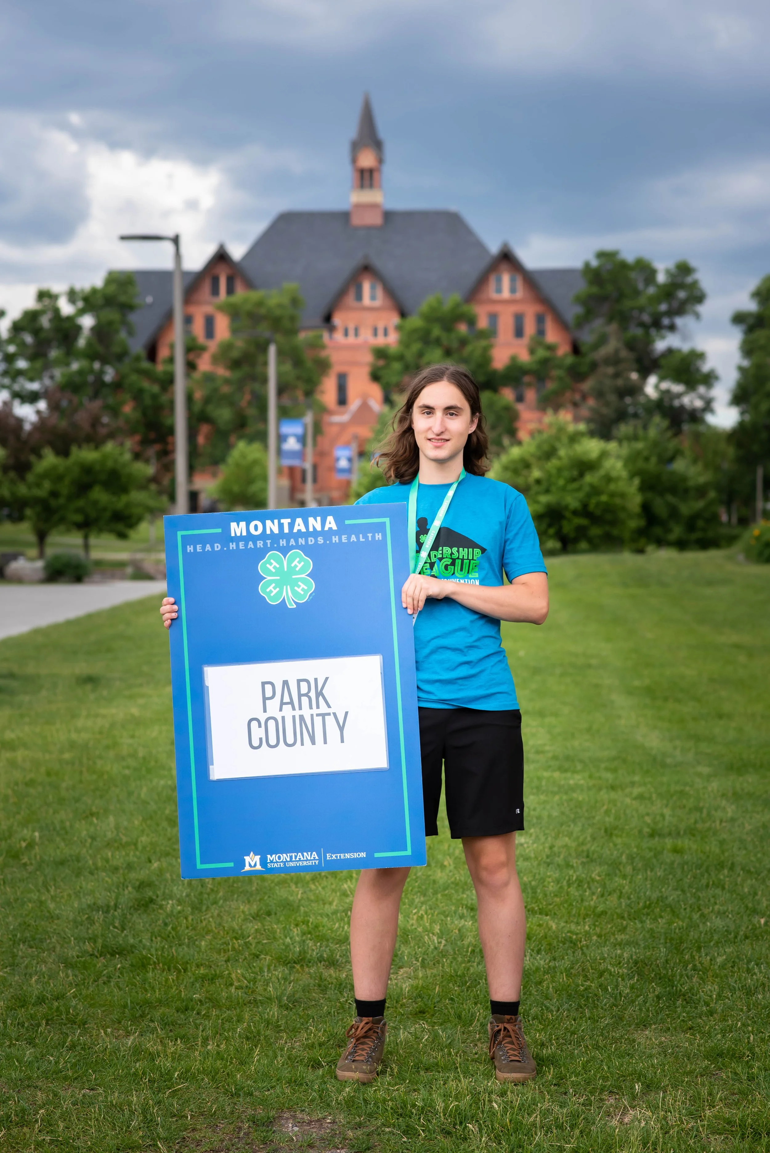 A young man with long brown hair wearing a blue T-shirt, black shorts, and brown hiking shoes standing outdoors on grass. He is holding a large blue sign that reads 'Park County' with the Montana State University logo. Behind him, there is a park with trees, pathways, and a historic brick building with a tall steeple under a cloudy sky.