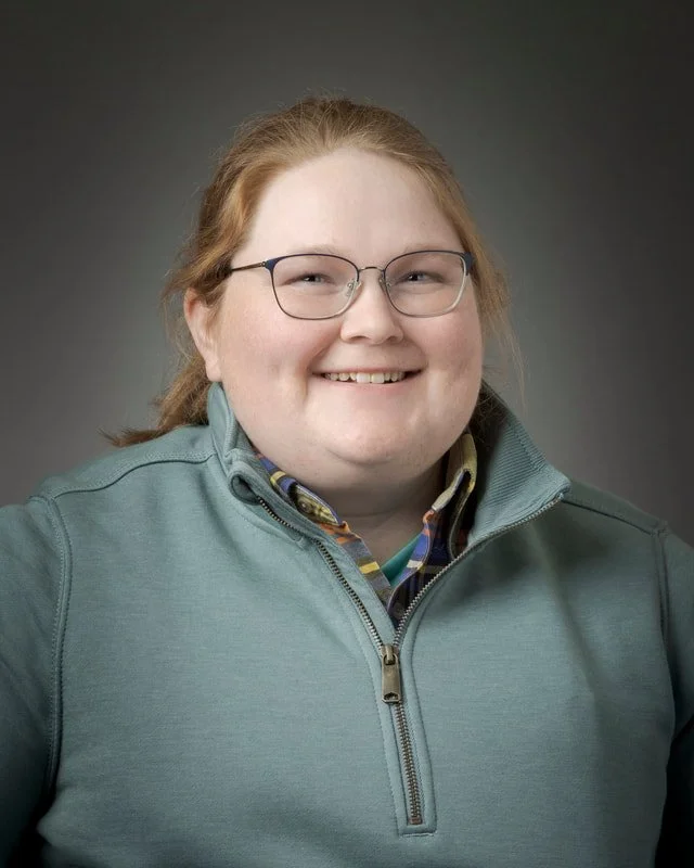 A young person with red hair and glasses smiling at the camera, wearing a teal jacket and a plaid shirt underneath.