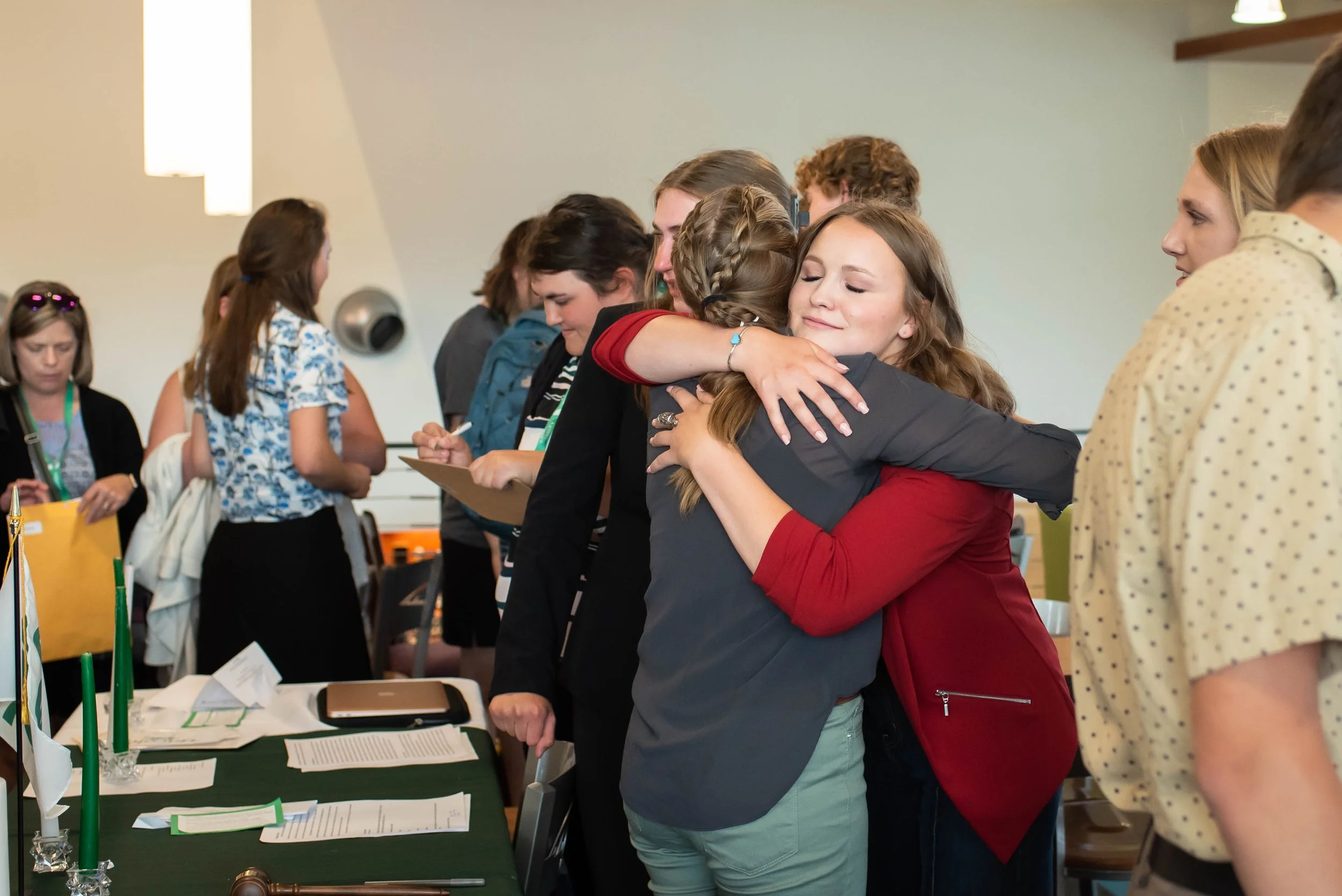 Two women hugging at a conference, with other attendees around them, some taking notes, in a room with tables and documents.