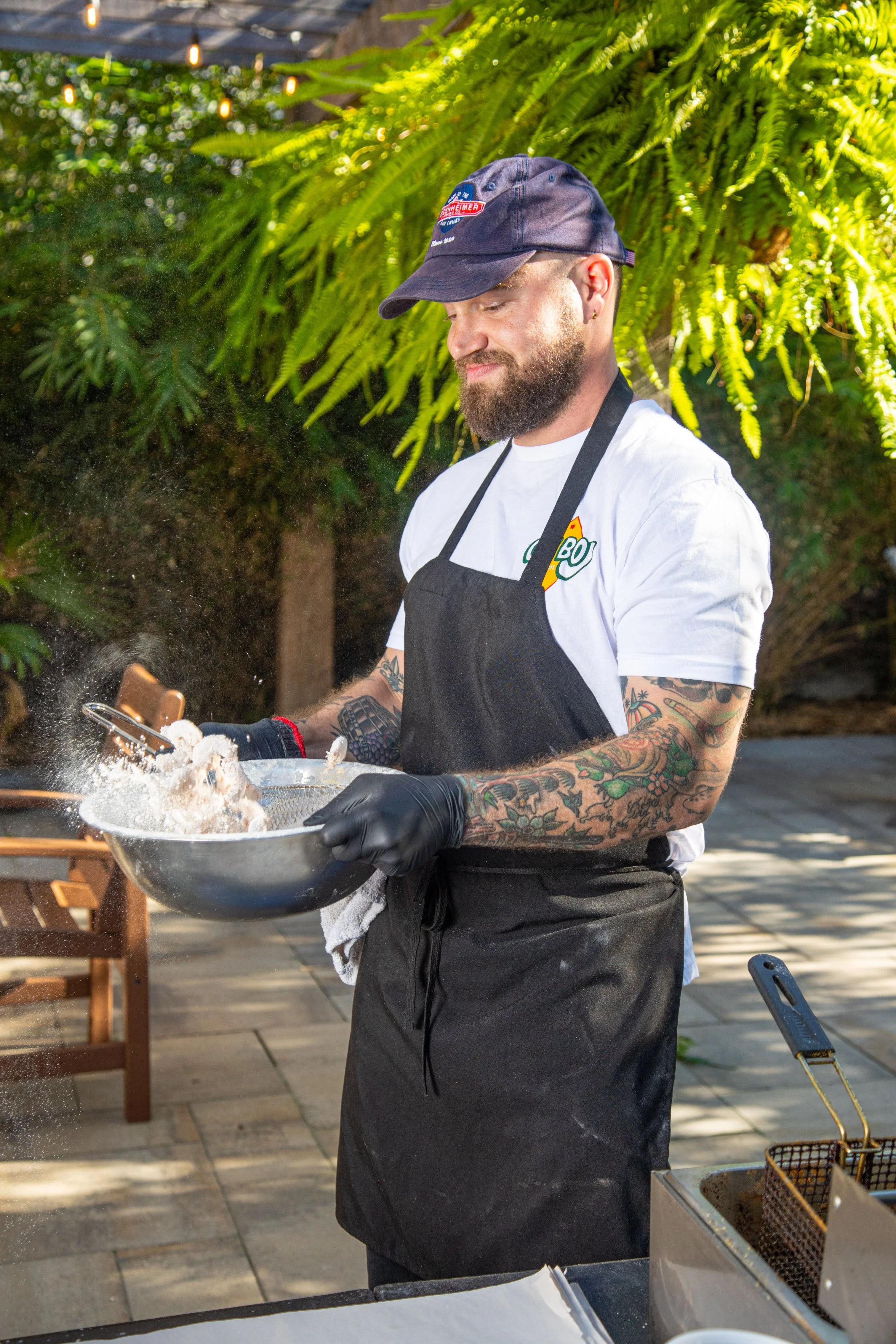 A man with tattoos, wearing a black apron, white T-shirt with a logo, a navy cap, and black gloves, is tossing food in a large metal bowl outdoors with lush green plants and hanging string lights in the background.