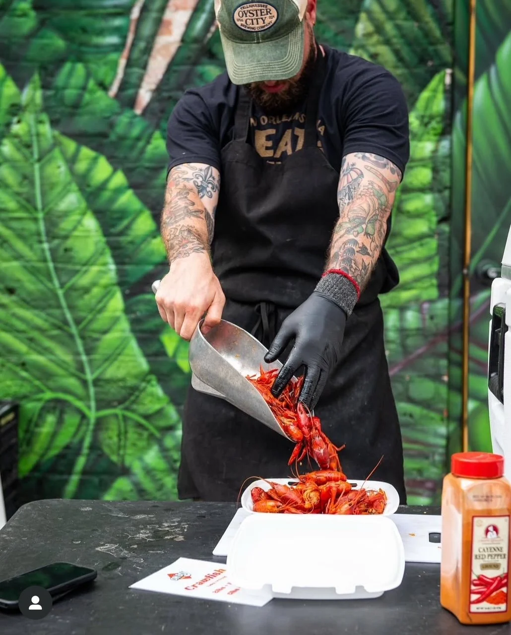 A man with tattoos wearing a cap, black T-shirt, and black gloves is pouring cooked crawfish from a metal scoop into a styrofoam container. There is a bottle of Red Pepper seasoning and a smartphone on the black table, with a leafy green jungle-themed backdrop behind him.