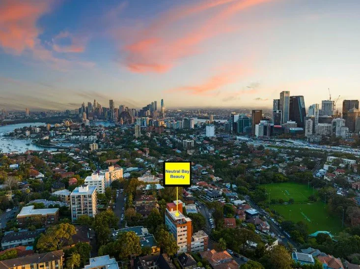Aerial view of a city skyline during sunset with a mix of high-rise buildings and residential areas, including a park. A yellow sign labeled 'Neutral Bay Beauty' is visible among the buildings.