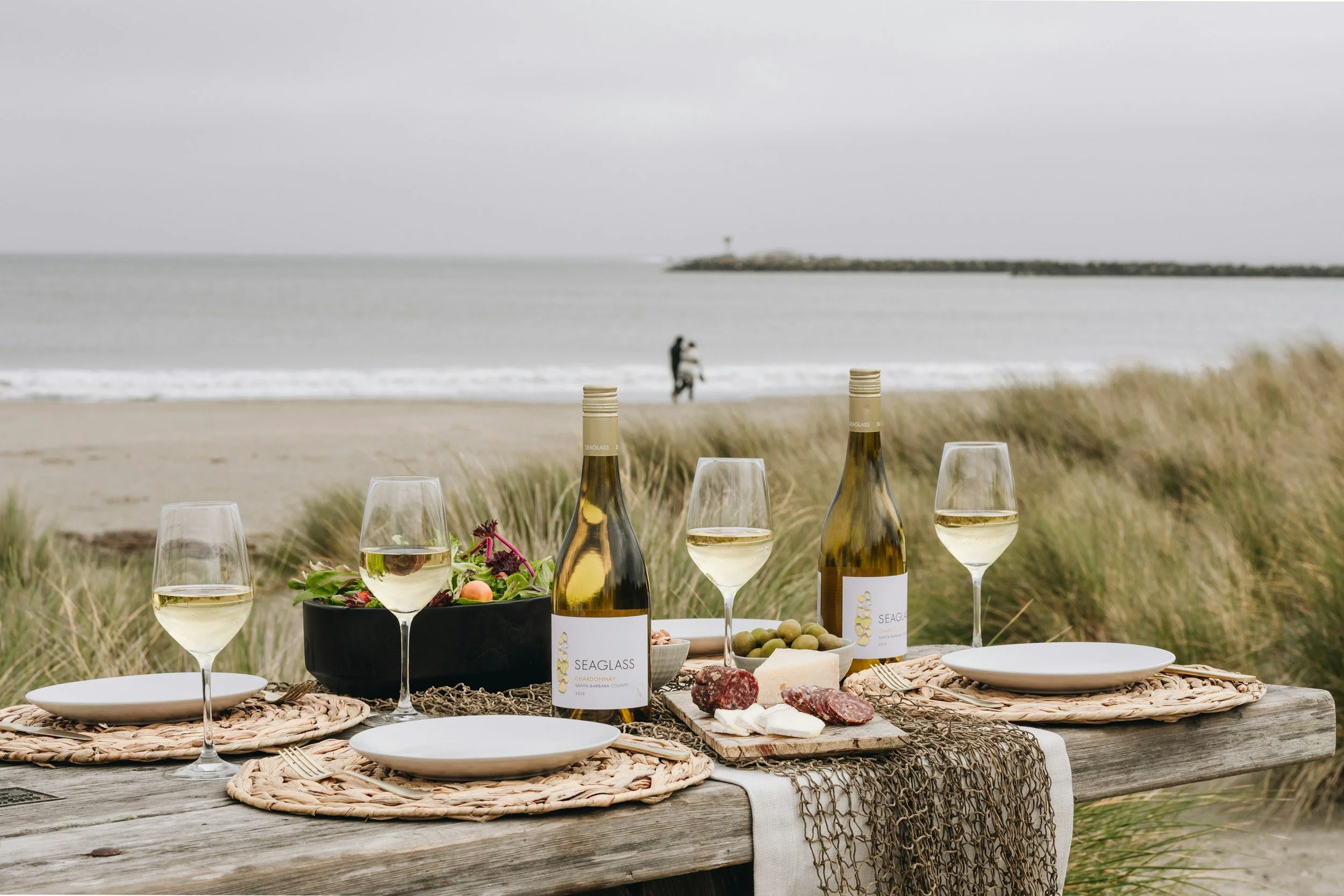 Beachside picnic setup with wooden table, bottles of wine, wine glasses, cheese, cured meats, charcuterie, salad, ocean and cloudy sky background. Nelson Bay, Port Stephens, Illawarra, Mollymook,  Sydney Northern Beaches, Jervis Bay, NSW South Coast