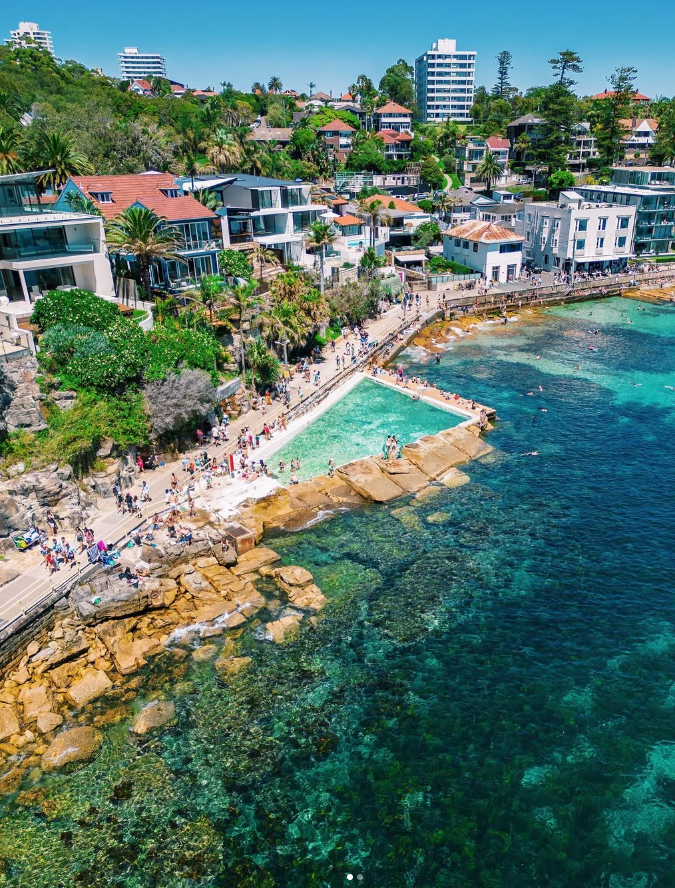 	
Coastal scene with rocky shoreline, ocean with clear blue water, and an ocean pool. Paved walkway with people. Modern residential buildings and lush greenery on a hillside. Palm Beach, Manly, Seaforth, Sydney Northern Beaches, Avalon, North Head.