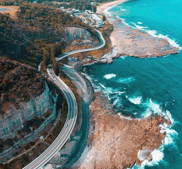 Winding coastal highway runs along a rocky cliffside next to ocean, with a beach and buildings in the background. Illawarra, NSW South Coast, Wollongong, Kiama, Berry, Gerringong, Gerroa, Hyams, Mollymook, Jervis Bay, Culburra.