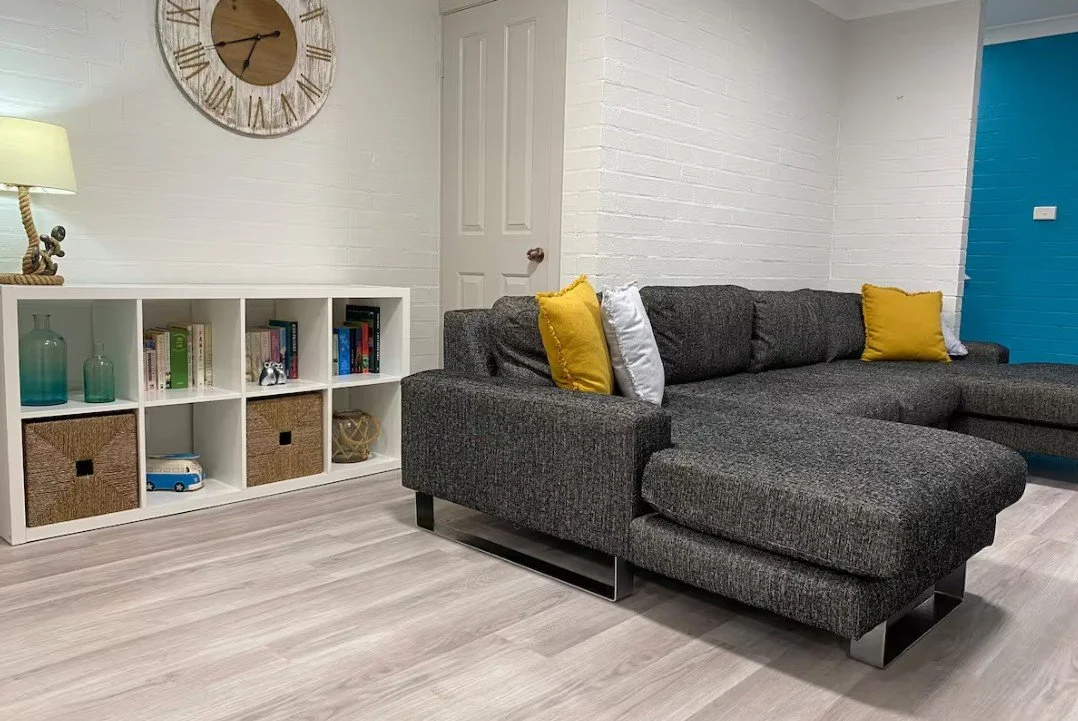 Living room with a dark gray sectional sofa with three cushions, two yellow and one white, against a white brick wall. To the left, a white bookshelf with decorative items, books, and storage baskets. A large wall clock with Roman numerals hangs above the bookshelf. A lamp with a beige shade and a nautical-themed base is on the bookshelf. To the right, a blue wall is partially visible.