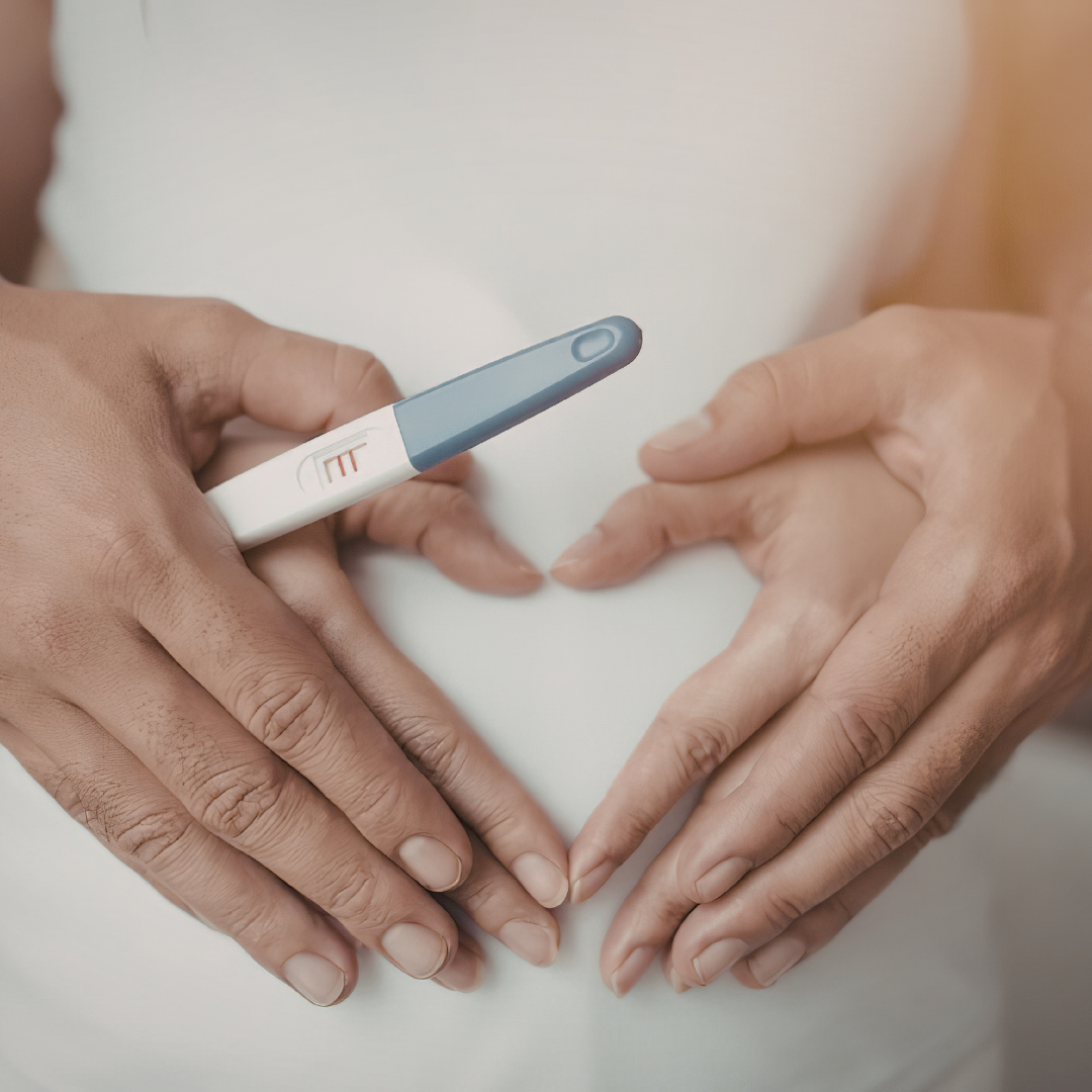 Two hands forming a heart shape with their fingers, and a positive pregnancy test held by one hand, with the test's display showing two lines.