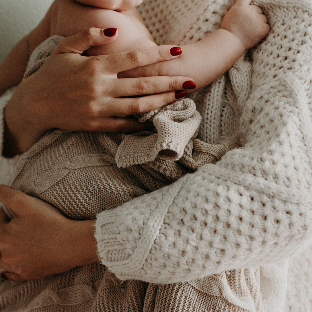 A mother holding her baby, gently supporting the baby's back with her hand. The baby is wrapped in a cozy, knitted blanket, and the mother is wearing a chunky, cream-colored knit sweater. The mother has red nail polish.