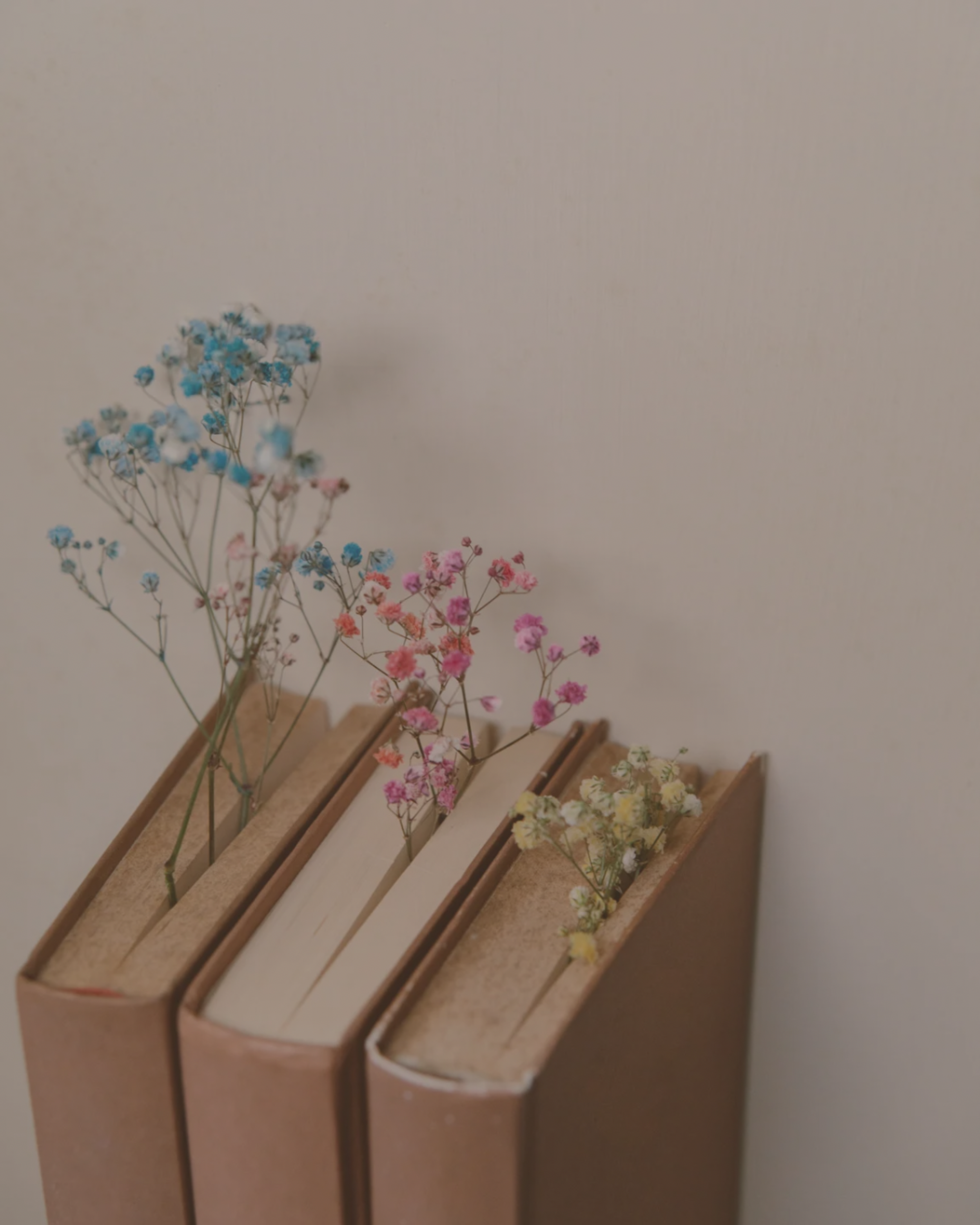 Three books with dried colorful flowers on top of their pages, arranged on a beige surface against a plain wall.