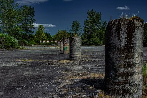 A row of weathered concrete pillars on a gravel lot with green trees and a blue sky in the background.