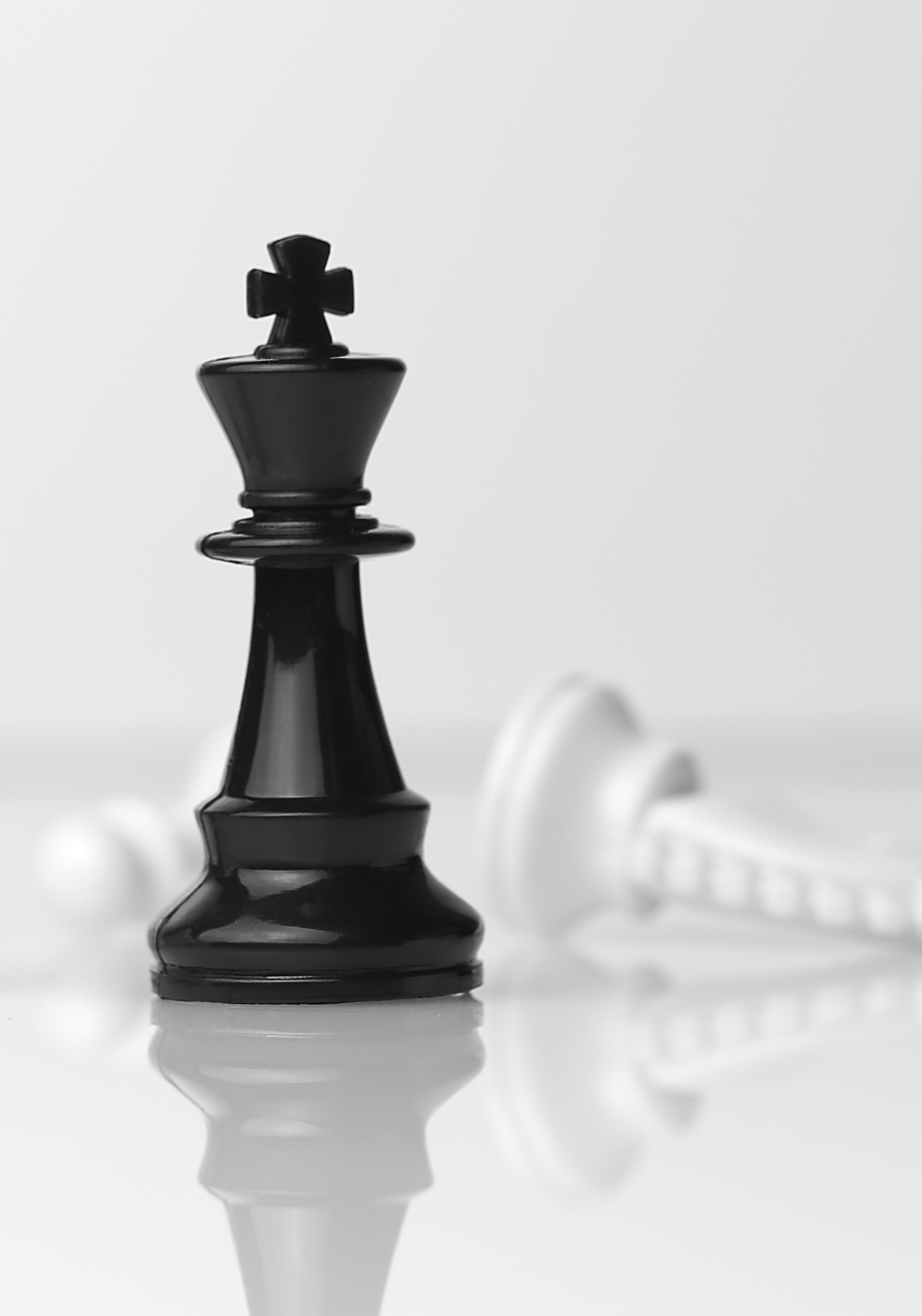 Black chess king standing upright on a reflective surface with a fallen white queen in the background.