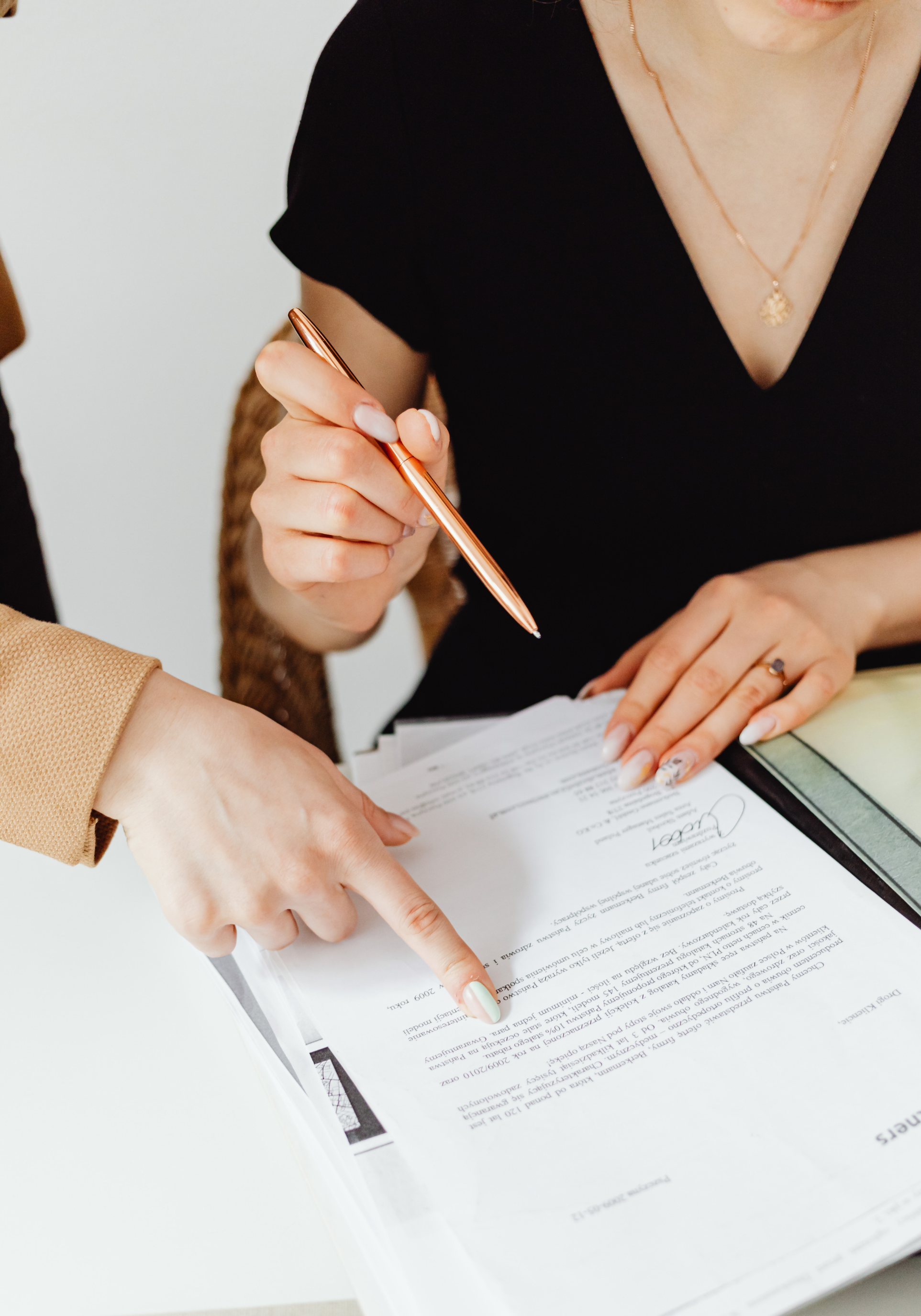 Two women reviewing and pointing at an outline of a book on a white desk, one holding a copper-colored pen.