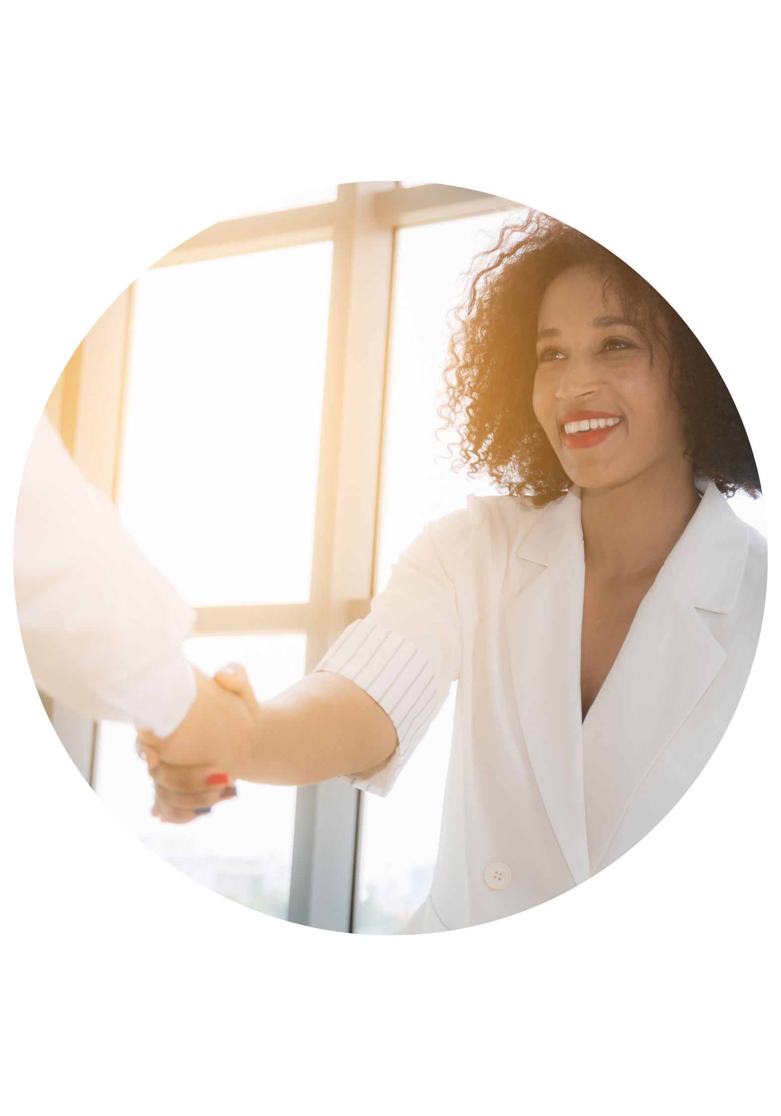 A woman with curly hair in a white blazer smiling and shaking hands with someone in a bright room with large windows.