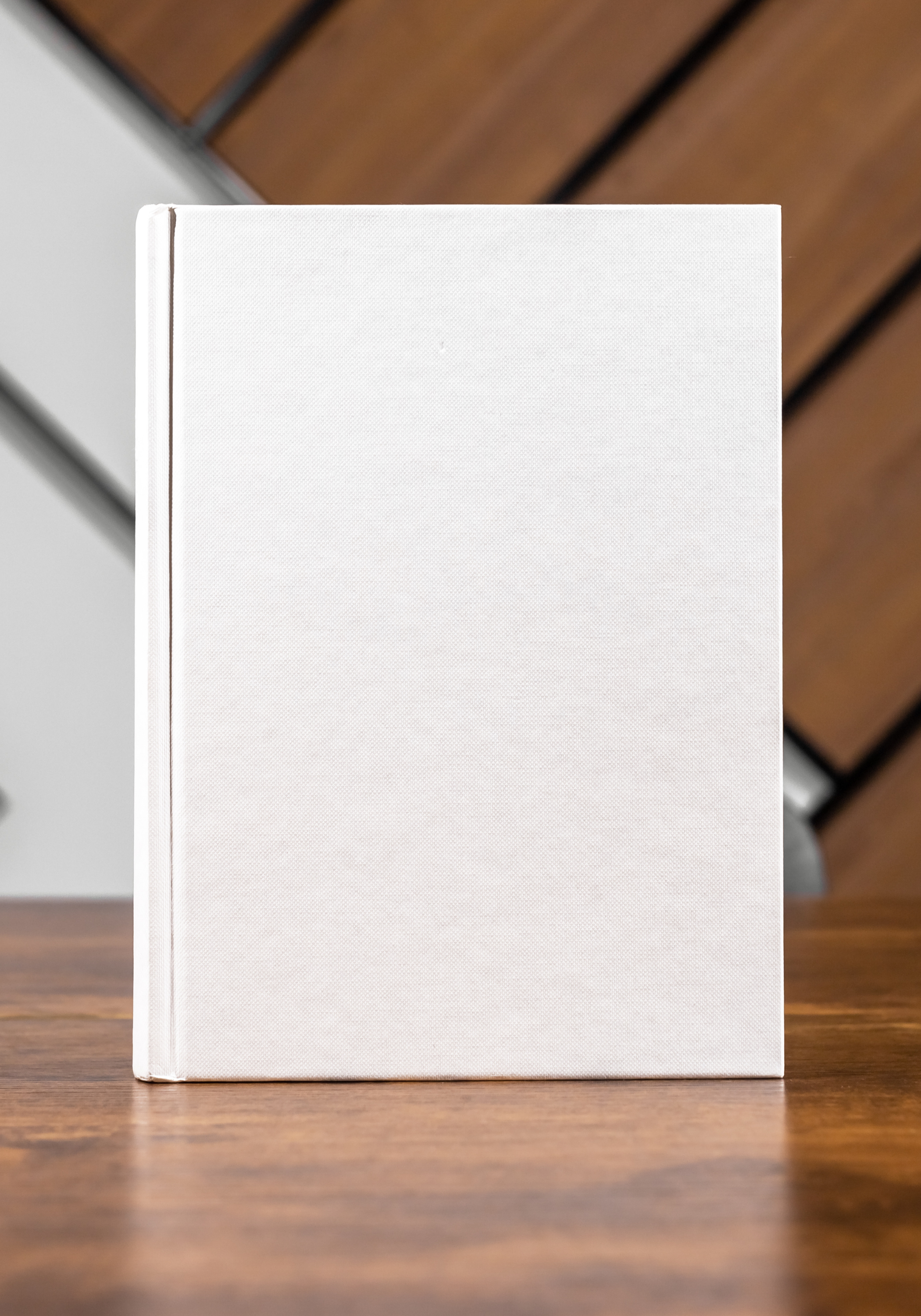A plain white hardcover book standing upright on a wooden surface with a wood-paneled wall and a gray cabinet in the background.