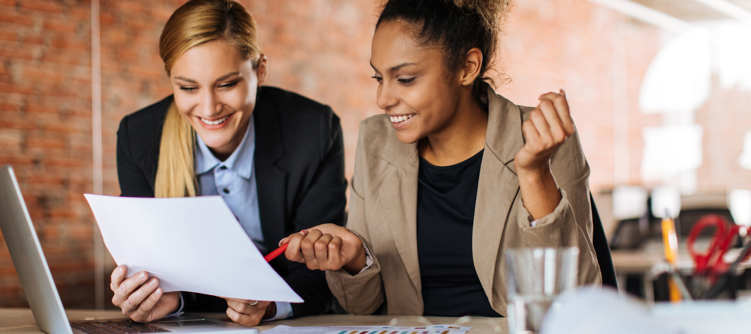 Two women are sitting at a table, smiling and looking at a paper and laptop in an office with a brick wall background.