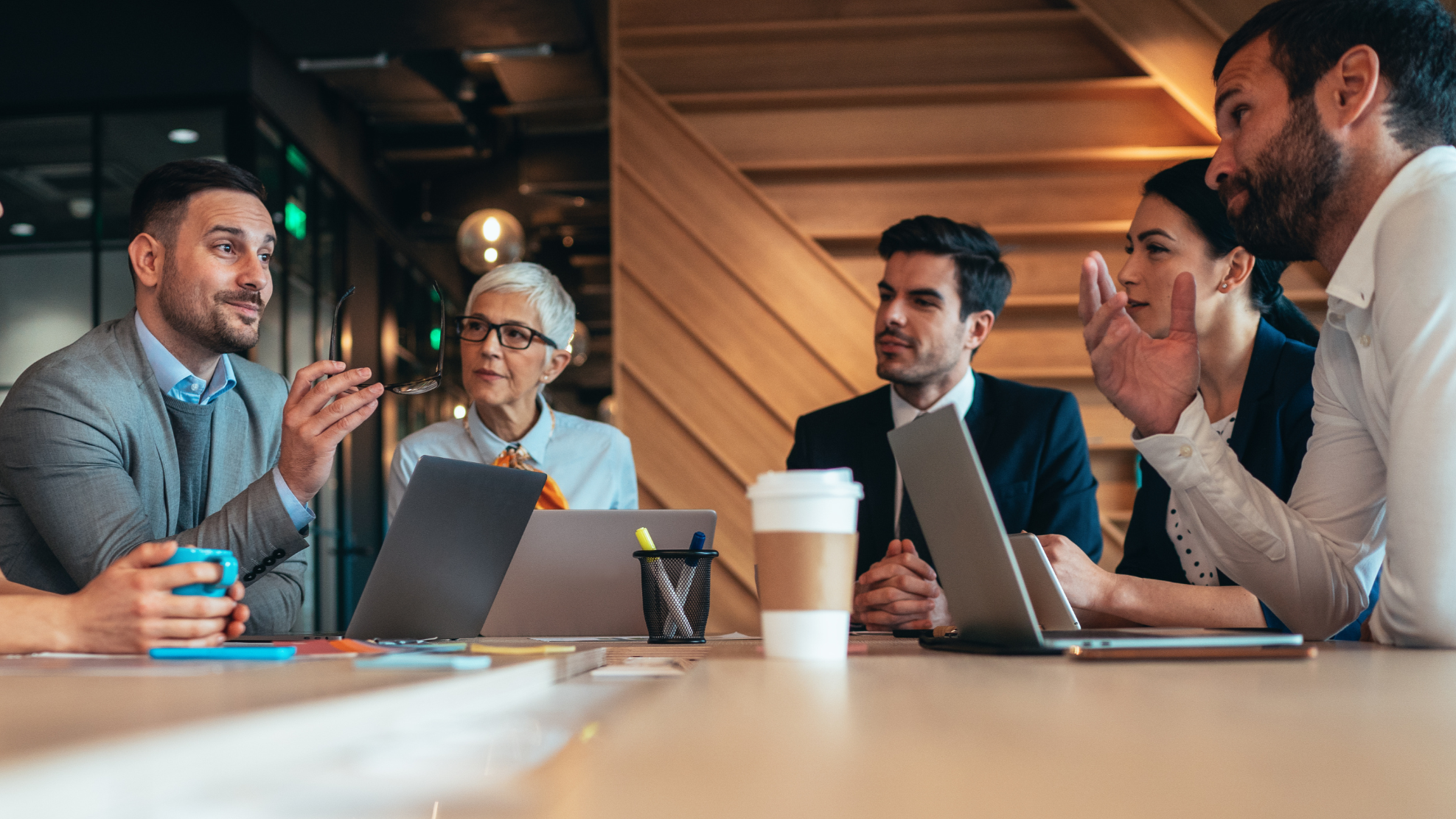 Business meeting with six people sitting around a table in a modern office or cafe, engaged in discussion, with laptops, notebooks, pens, and a coffee cup on the table.