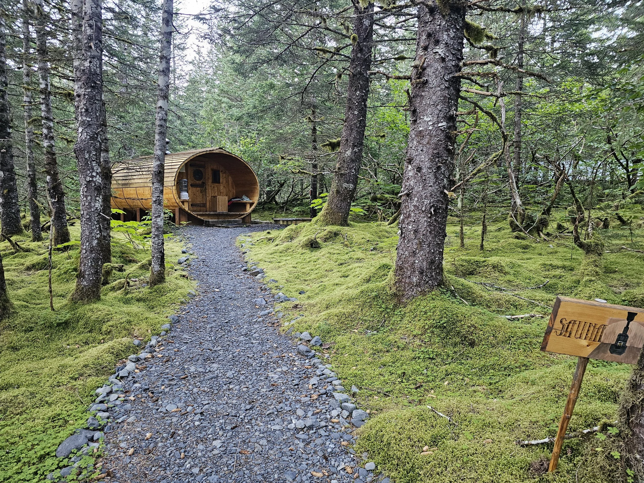 Barrel-shaped wooden sauna at Kenai Fjords Glacier Lodge, set along a gravel path in a moss-covered evergreen forest.