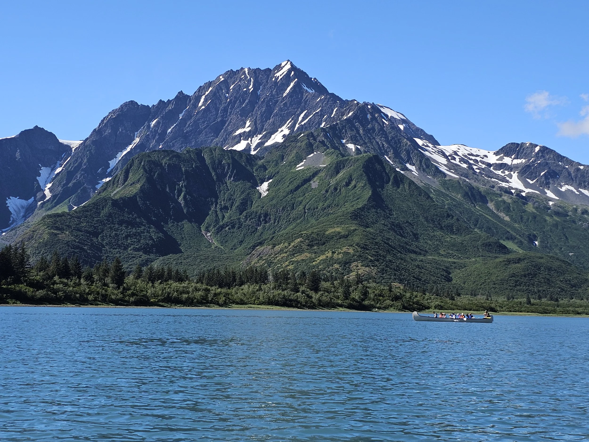 Canoe crossing calm blue water in Pedersen Lagoon, Alaska, with forested shoreline and tall snow-streaked mountains in the background.