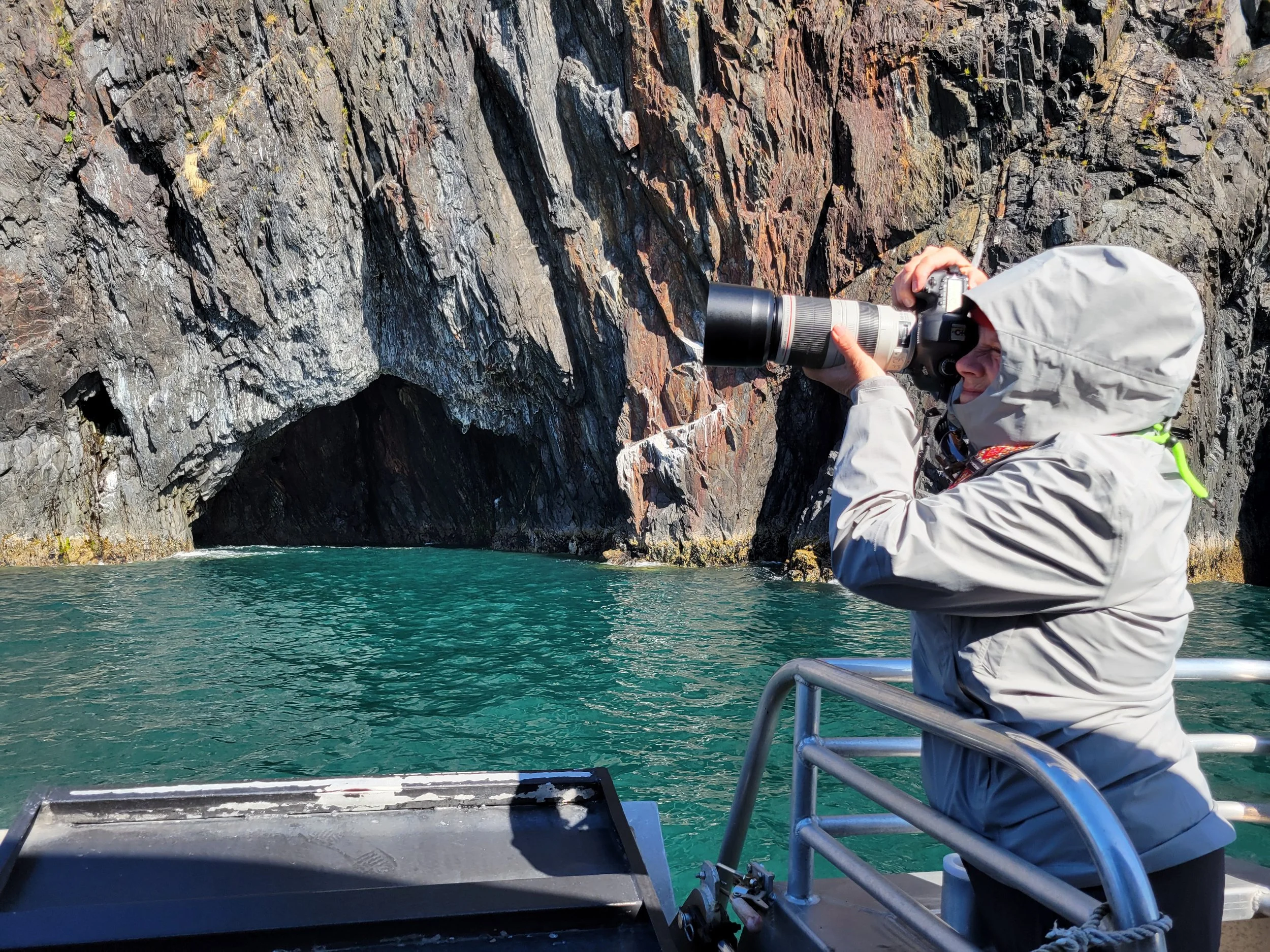 Danielle photographing steep rocky cliffs from the deck of a boat in Kenai Fjords National Park, Alaska, with turquoise water below.
