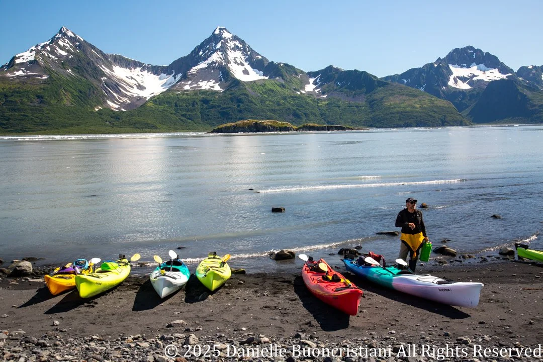 Colorful sea kayaks lined up on a rocky beach in Aialik Bay, Kenai Fjords National Park, with snow-covered mountains in the background.