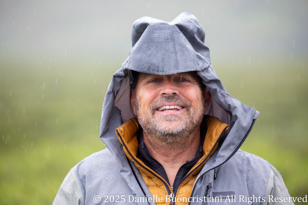 Portrait of a man in a rain jacket standing in steady rain in Denali National Park, water droplets visible as stormy weather continues.