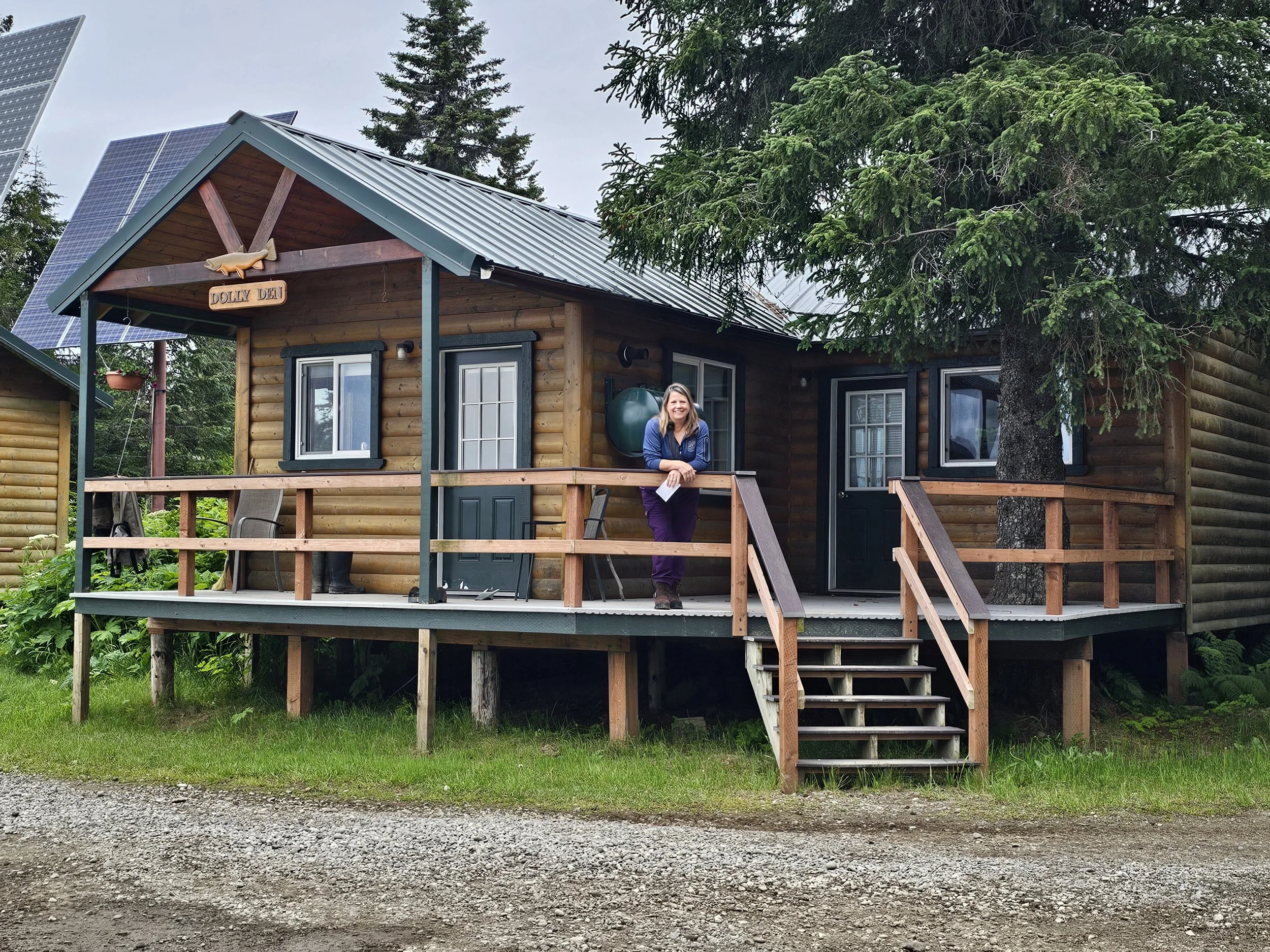 A log cabin at Silver Salmon Creek Lodge with Danielle standing on the front deck, surrounded by trees and solar panels.