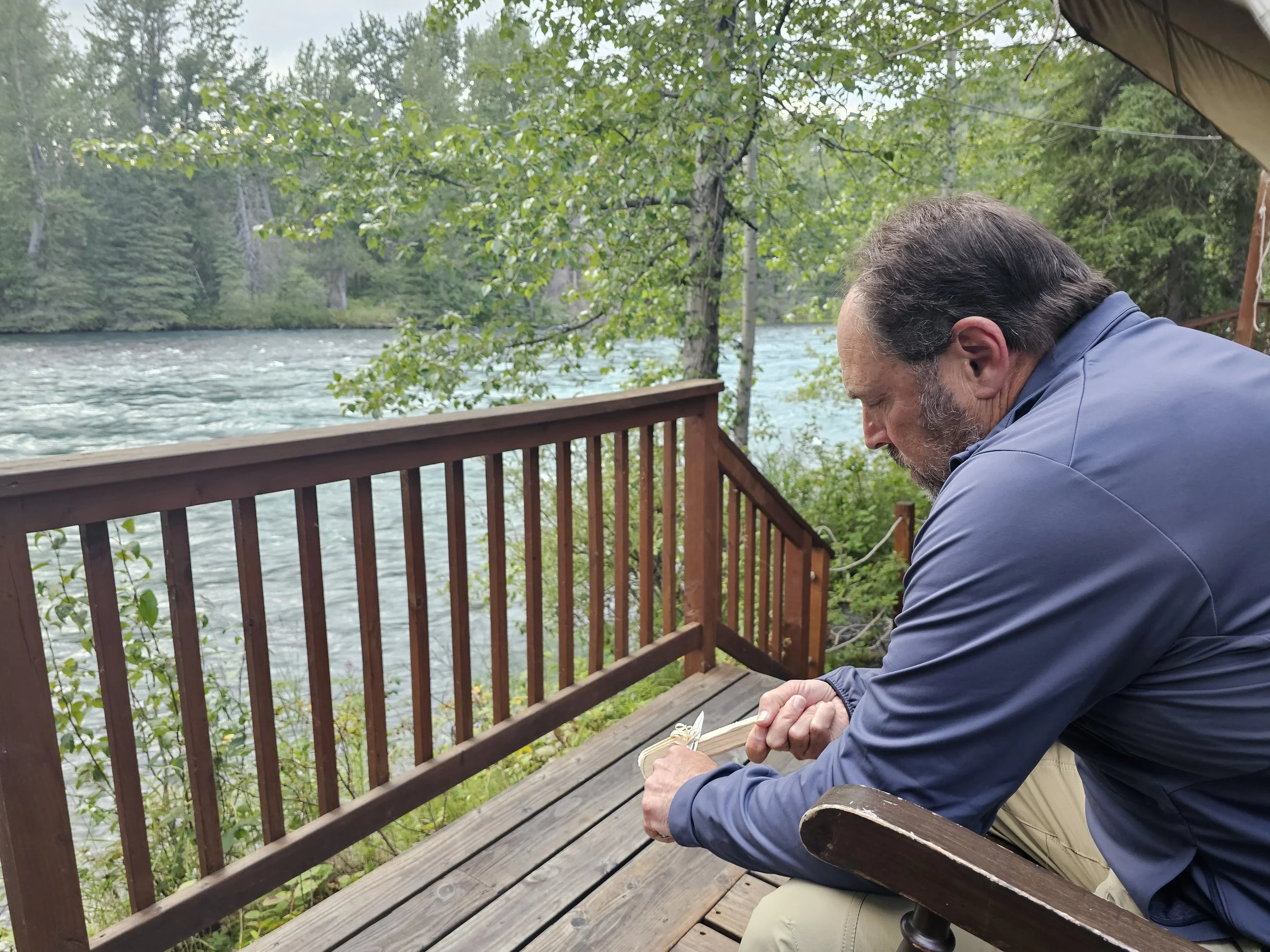 Dave sitting in a wooden chair carving a piece of wood beside the flowing Kenai River, with trees lining the opposite bank.