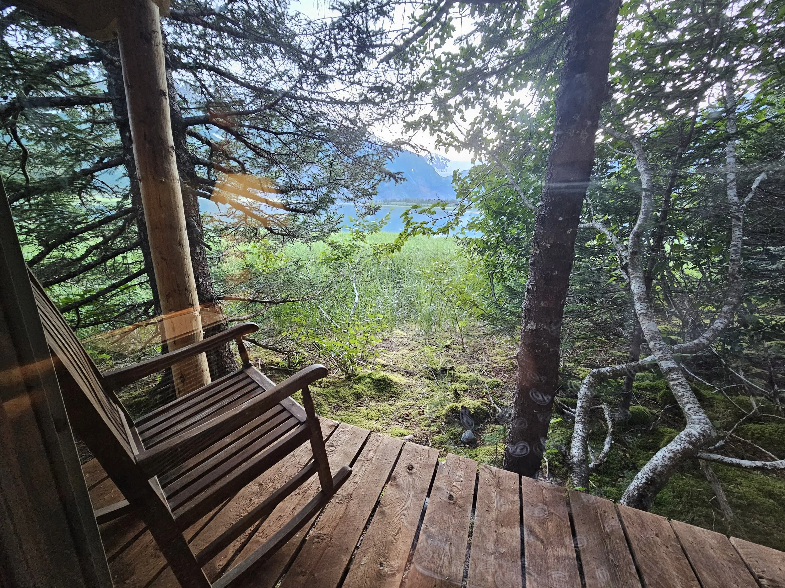 Wooden rocking chair on a private cabin porch at Kenai Fjords Glacier Lodge, looking through evergreen trees toward Pedersen Lagoon and the mountains beyond.