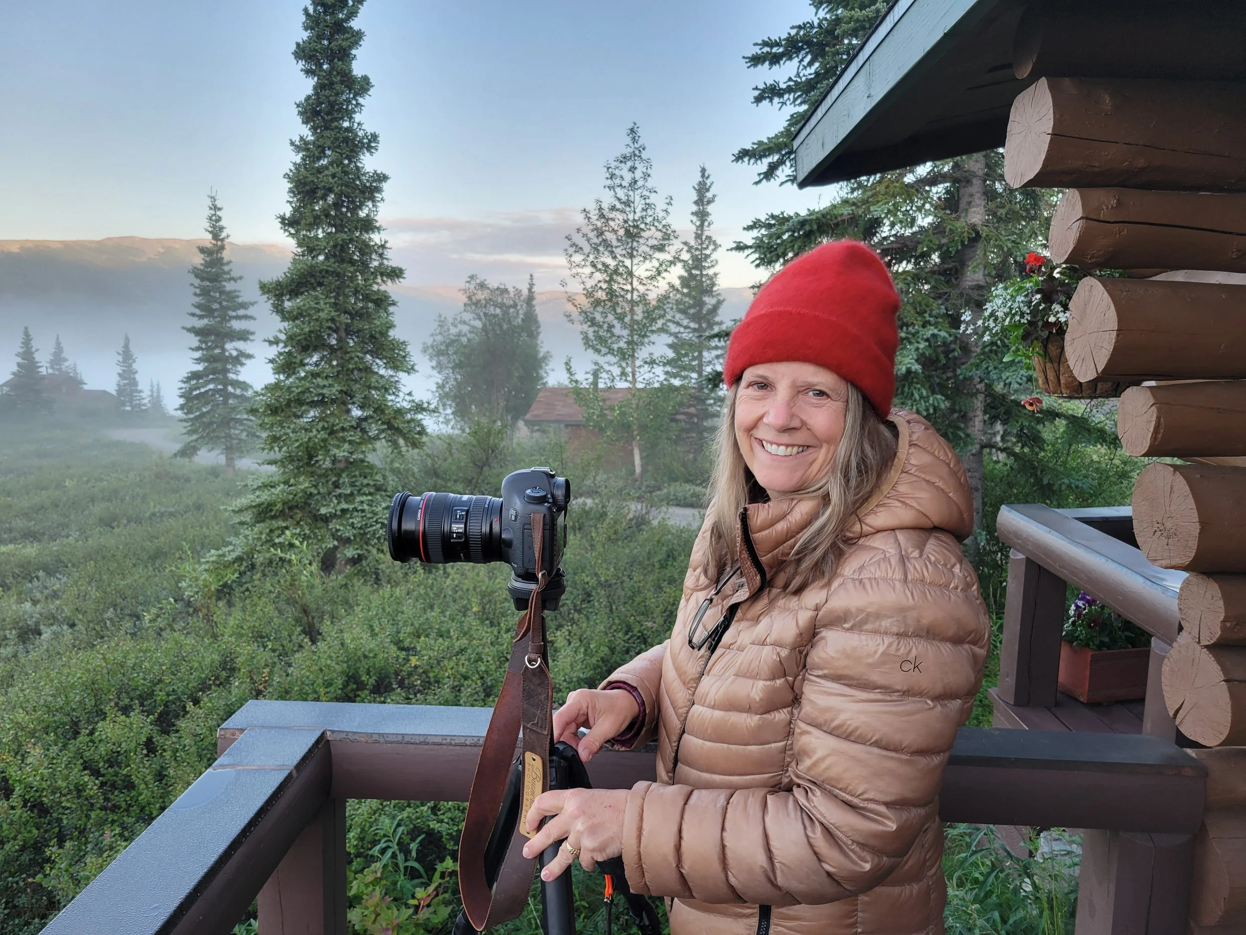 Photographer smiling beside a camera on a tripod on a lodge deck at Camp Denali, with mist in the valley and early morning light in the background.