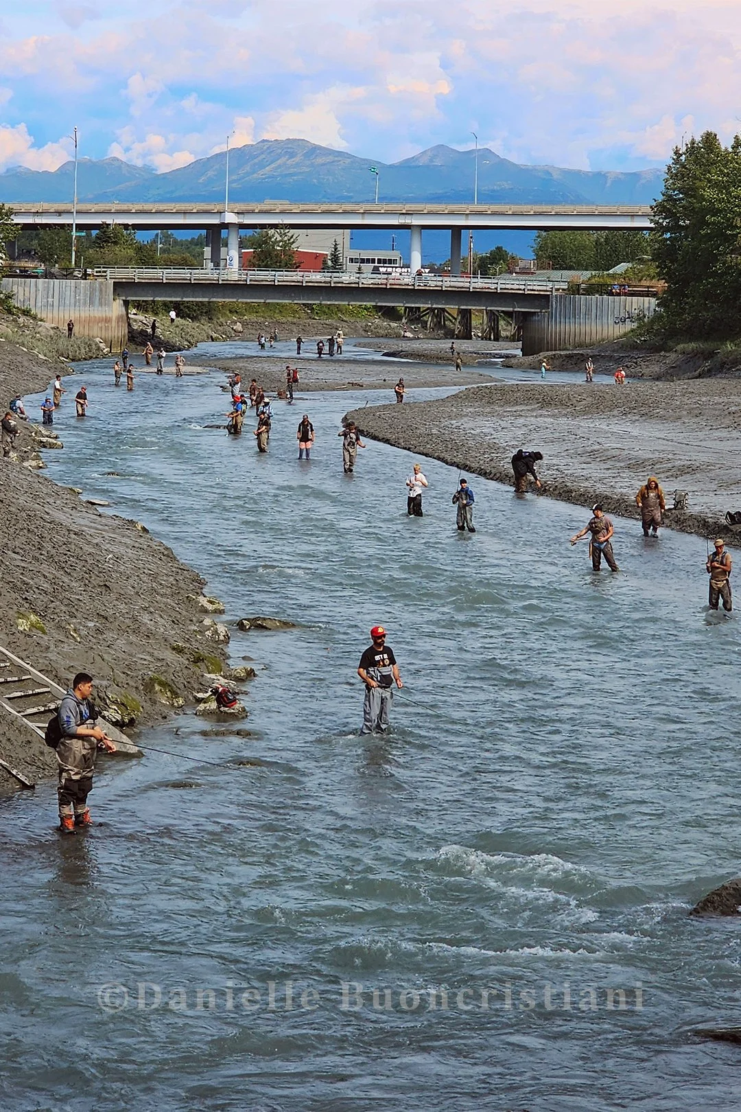 People fishing for salmon in Ship Creek in Anchorage, Alaska, during a salmon derby, with bridges and mountains in the background.