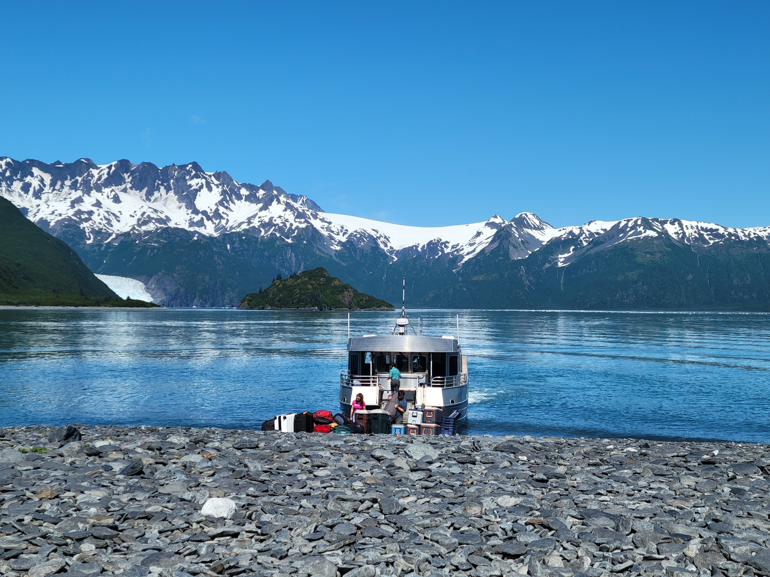 Small boat pulled onto a rocky shoreline in Kenai Fjords National Park, Alaska, with snow-covered mountains and calm blue water in the background.