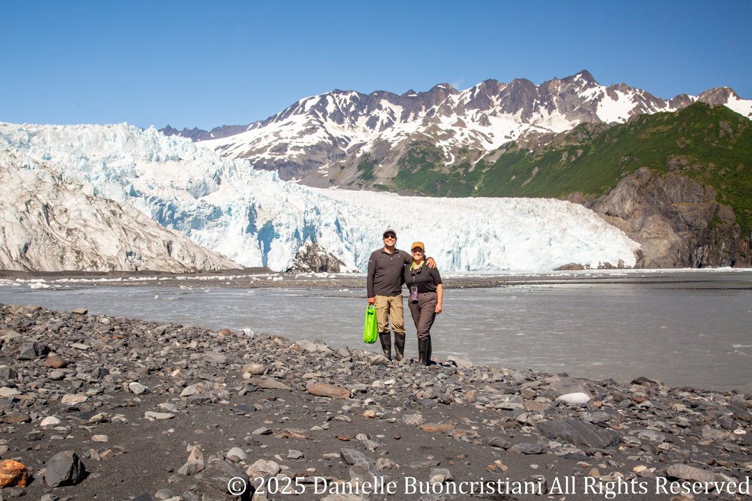 Couple standing on rocky shoreline in front of Aialik Glacier in Kenai Fjords National Park, Alaska.
