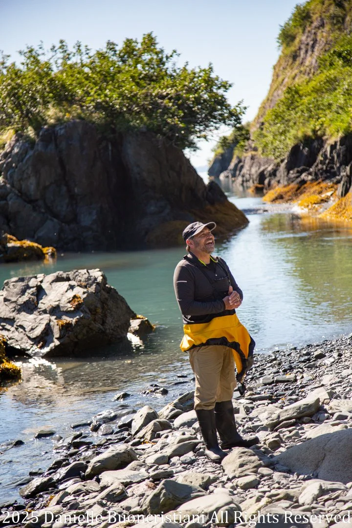 Man standing on a rocky shoreline in Aialik Bay, Kenai Fjords National Park, with steep coastal cliffs and calm water behind him.