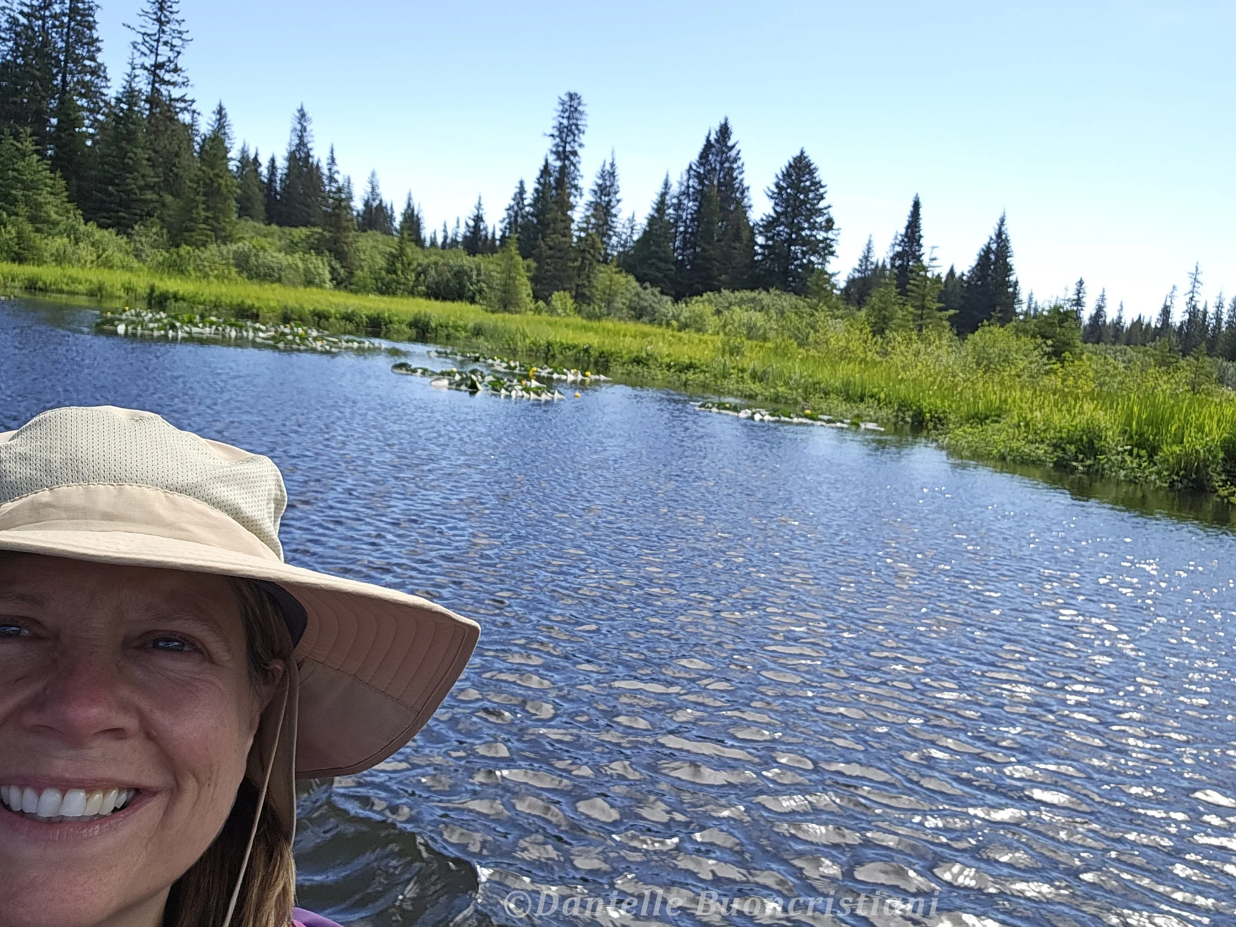 Photographer standing beside a calm lake near Silver Salmon Creek Lodge, with lily pads on the water and spruce trees along the shoreline.