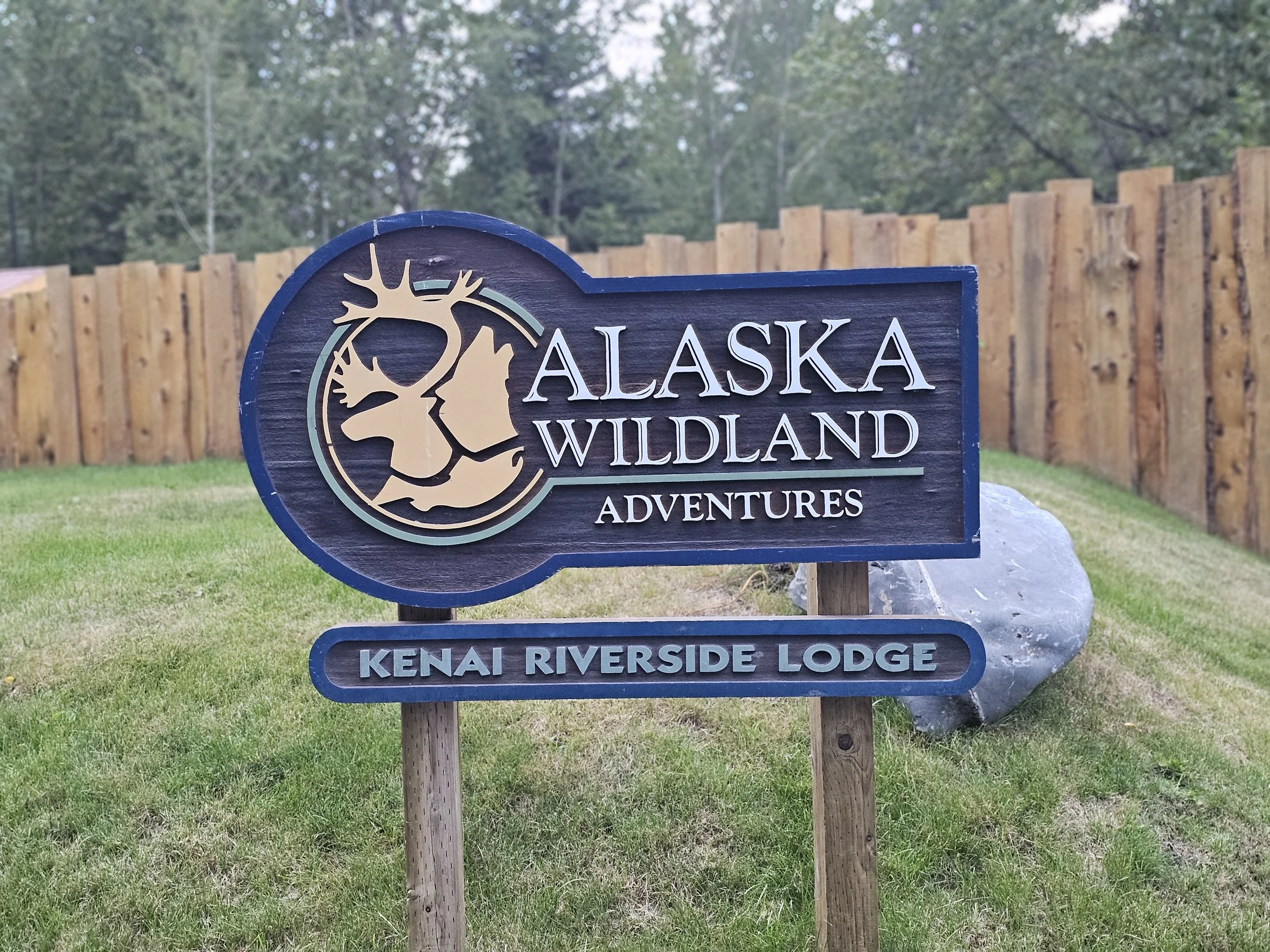 Wooden Alaska Wildland Adventures sign for Kenai Riverside Lodge beside a grassy path and tall wooden fence.