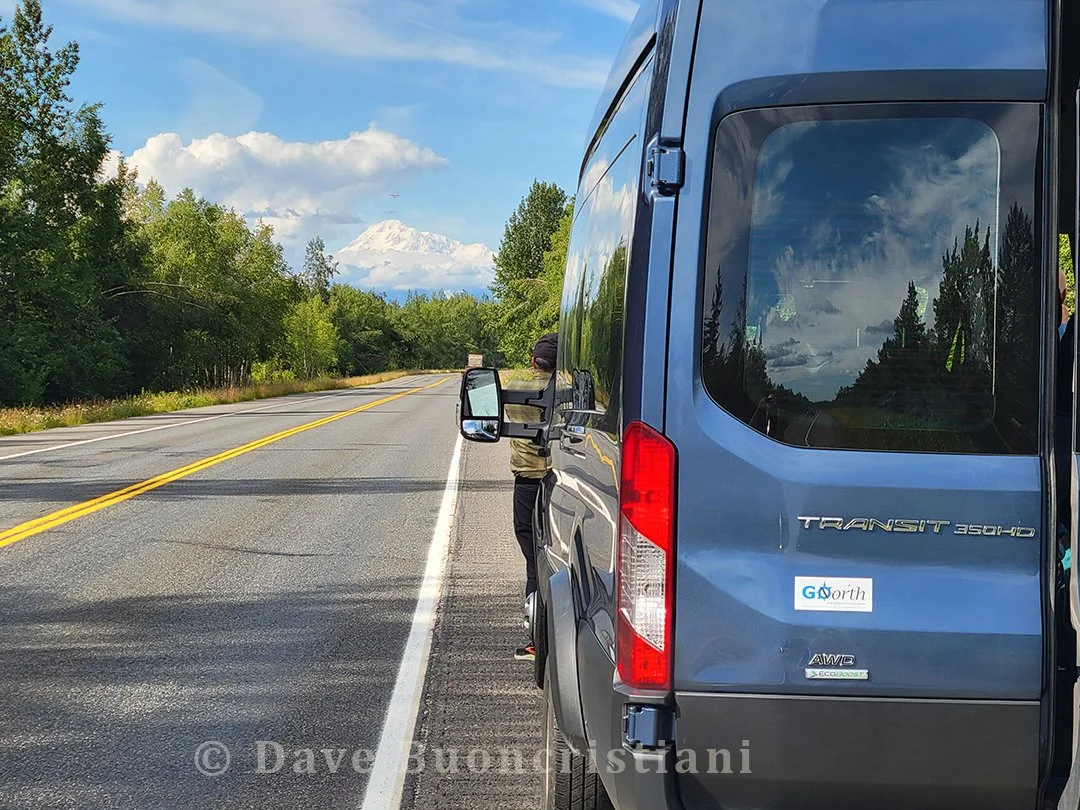Passenger van pulled over along an Alaska highway with Denali visible in the distance through breaks in the clouds.