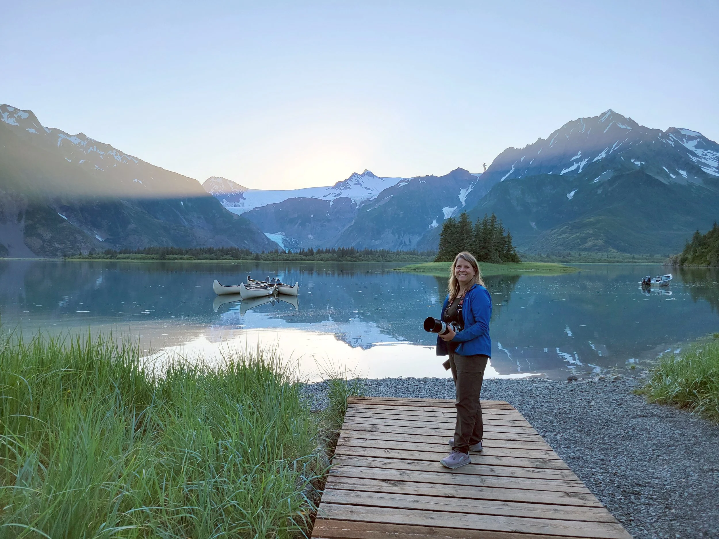 Danielle standing near Pedersen Lagoon in Alaska holding a camera, with calm water, canoes, and snow-capped mountains in the background during soft evening light.