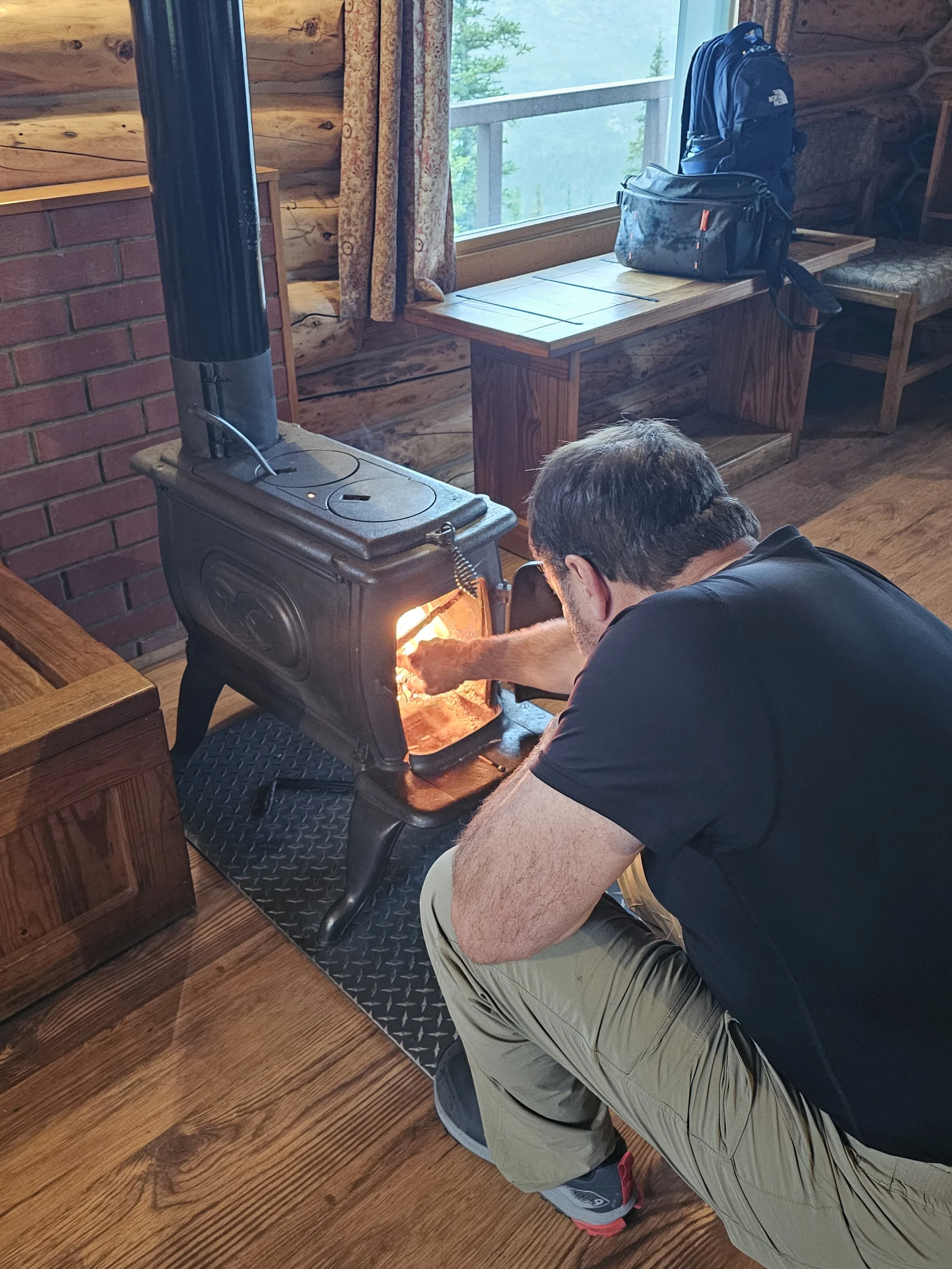 Man lighting a wood stove inside a rustic cabin at Camp Denali during rainy weather.