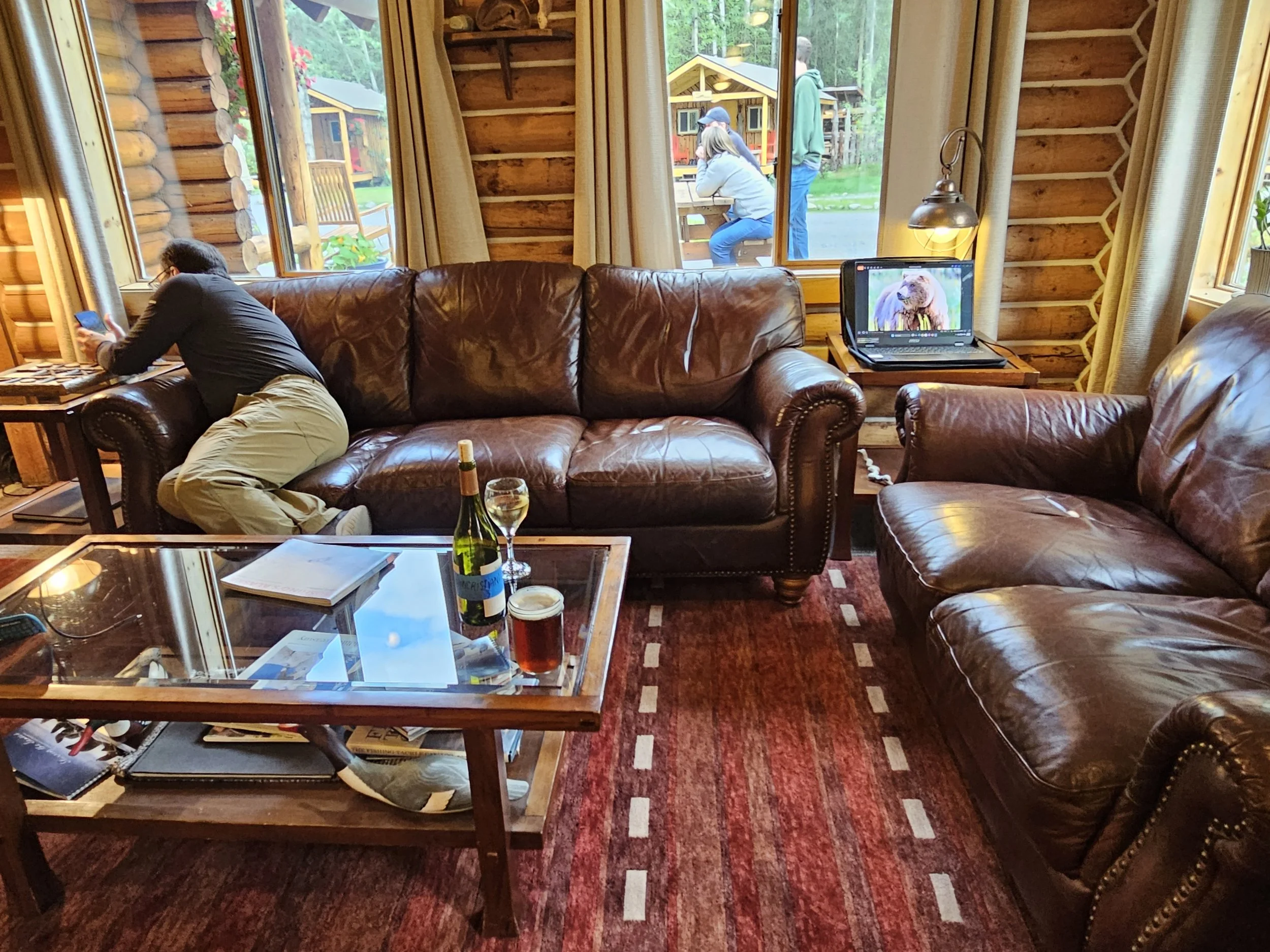 Interior lounge at Kenai Riverside Lodge with leather sofas, log walls, a glass coffee table, and an open laptop displaying a bear photograph.