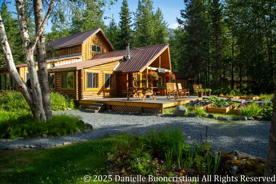 Main lodge building at Kenai Riverside Lodge in Alaska with wooden deck, chairs, and garden beds surrounded by forest.