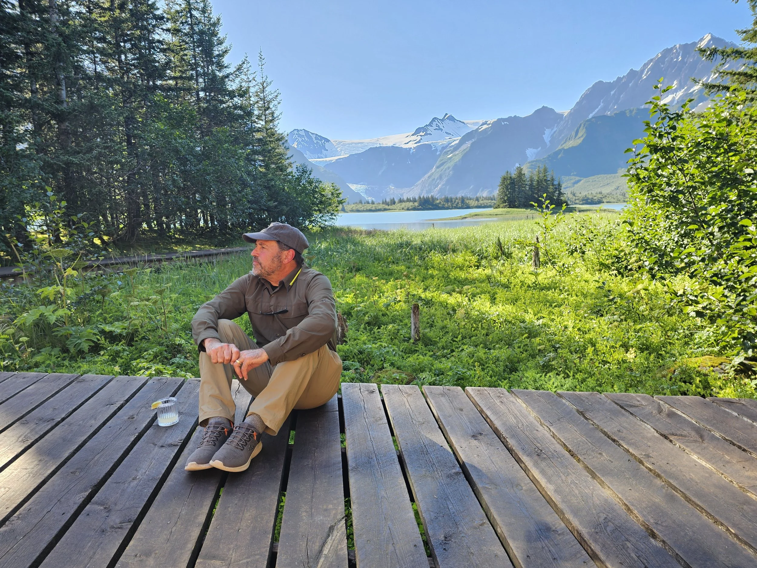 Man sitting on a wooden deck at Kenai Fjords Glacier Lodge, Alaska, looking toward Pedersen Lagoon and surrounding mountains in evening light.