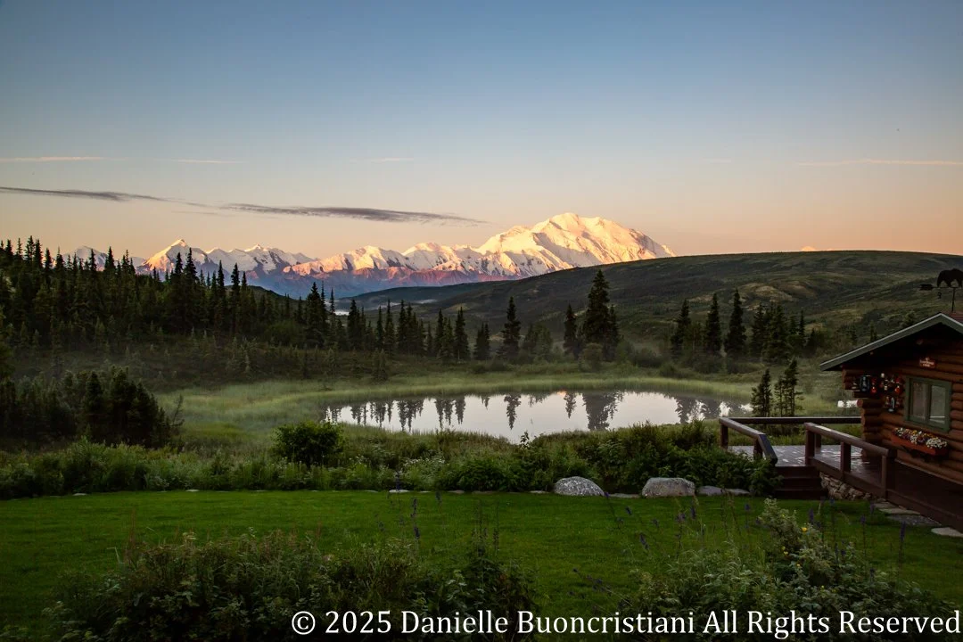 Sunrise light illuminates Mount Denali as it emerges from clouds, reflected in a pond near Camp Denali in Denali National Park.