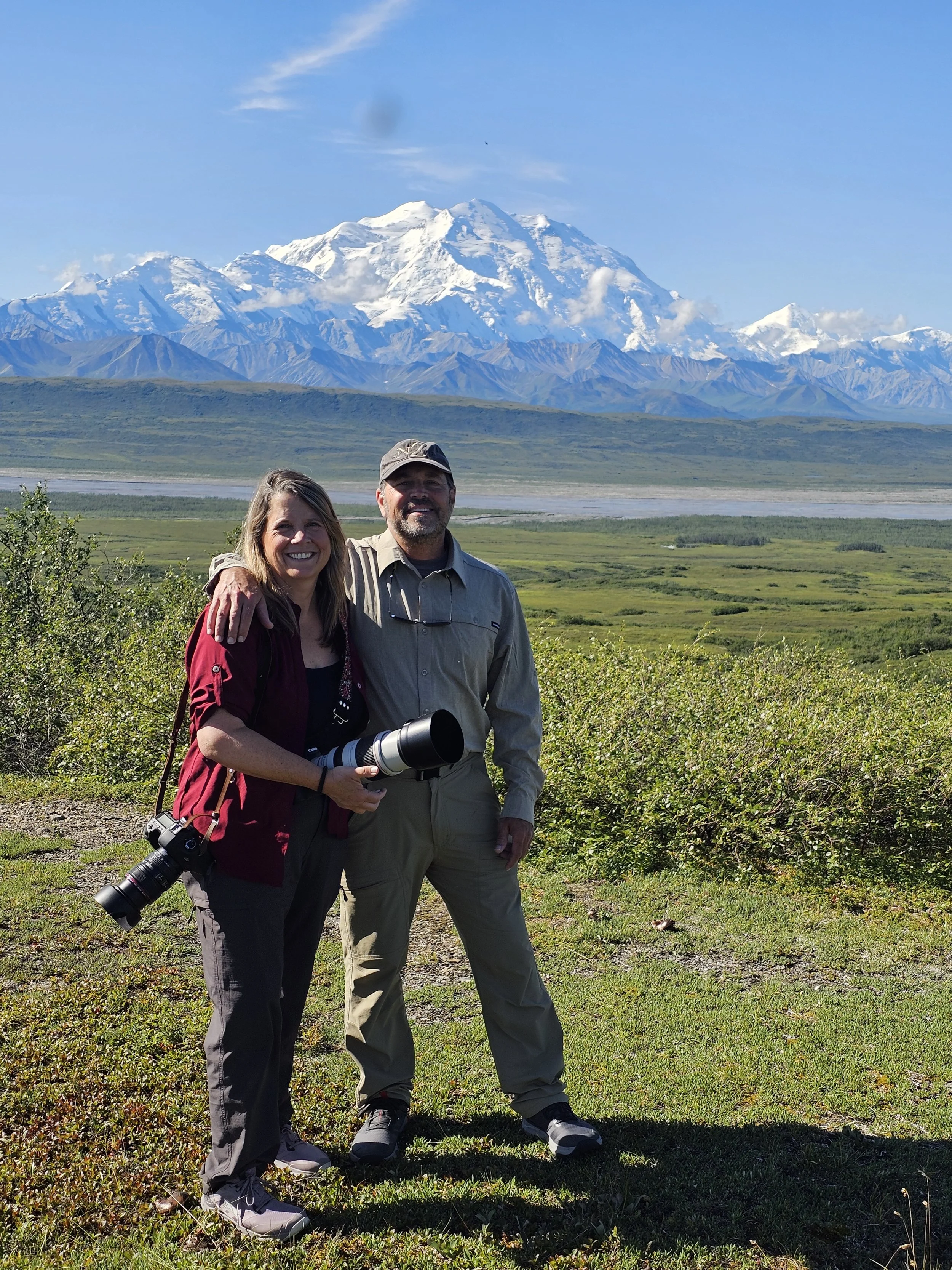 Danielle Buoncristiani and her husband Dave standing in Denali National Park holding cameras, smiling with Mount Denali in the background.