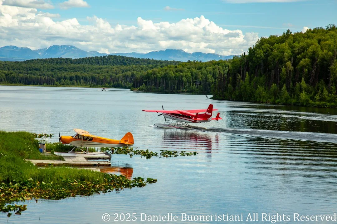 Two floatplanes on a calm lake in Alaska, with forested shoreline and distant mountains under a partly cloudy sky.