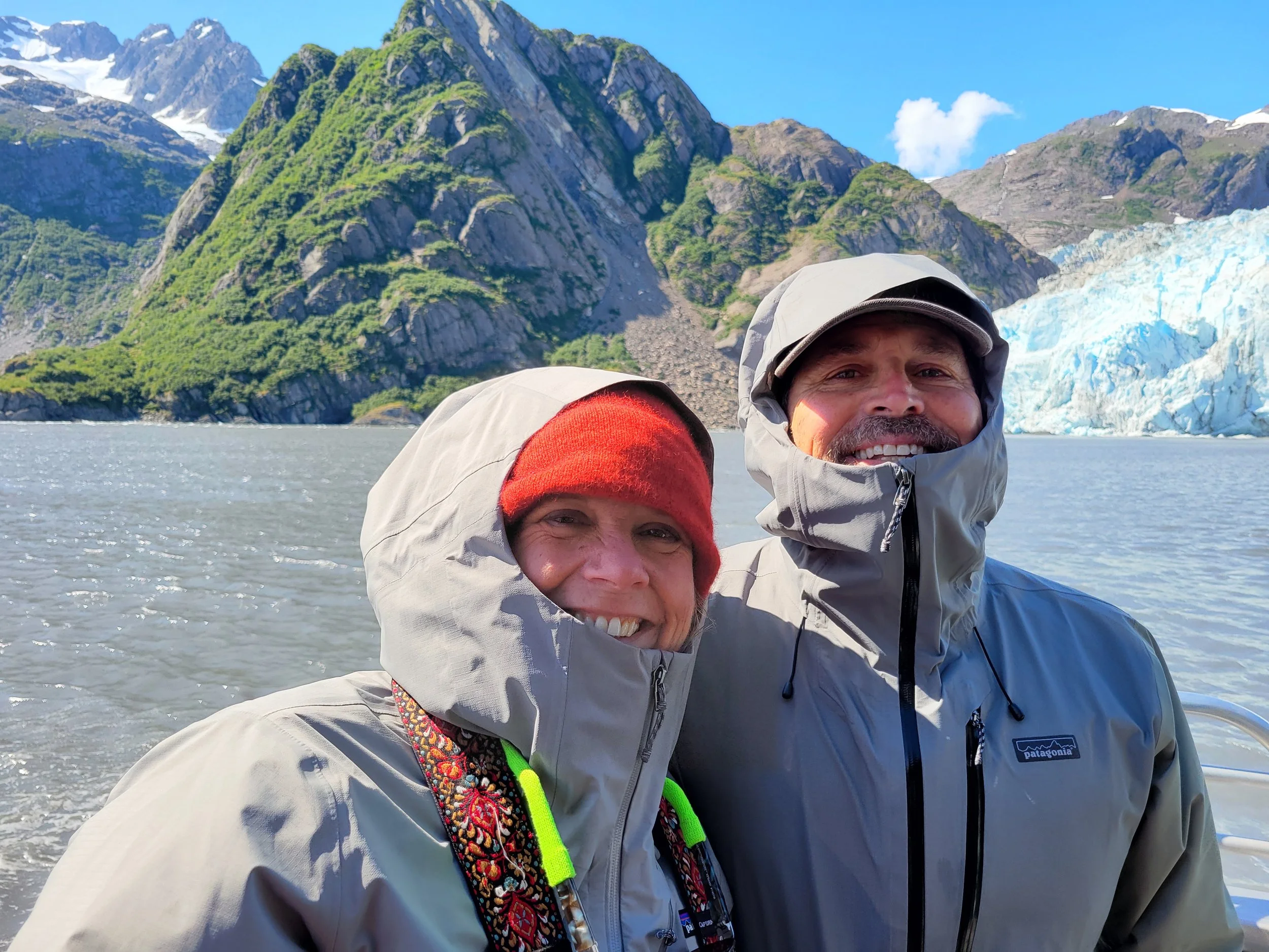 Danielle and Dave standing on a boat in Kenai Fjords National Park, Alaska, bundled in jackets with a blue glacier and steep mountains behind them.