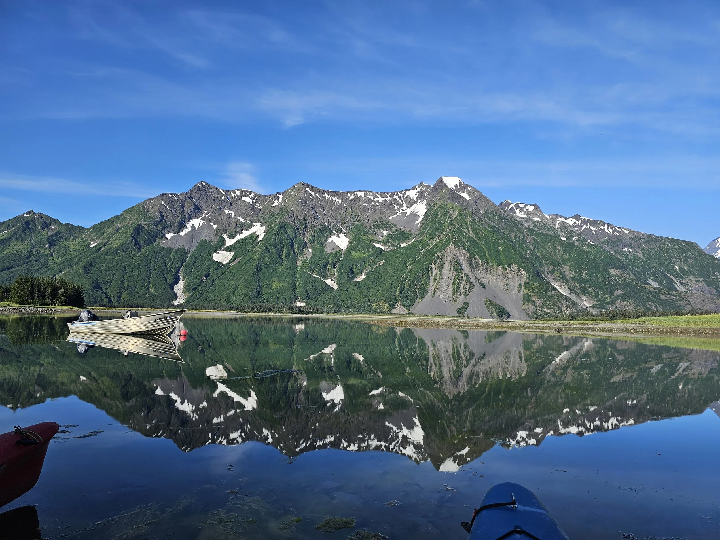 Calm water at Pedersen Lagoon reflecting green mountains with patches of snow, with small boats and kayaks near the shore at Kenai Fjords Glacier Lodge.
