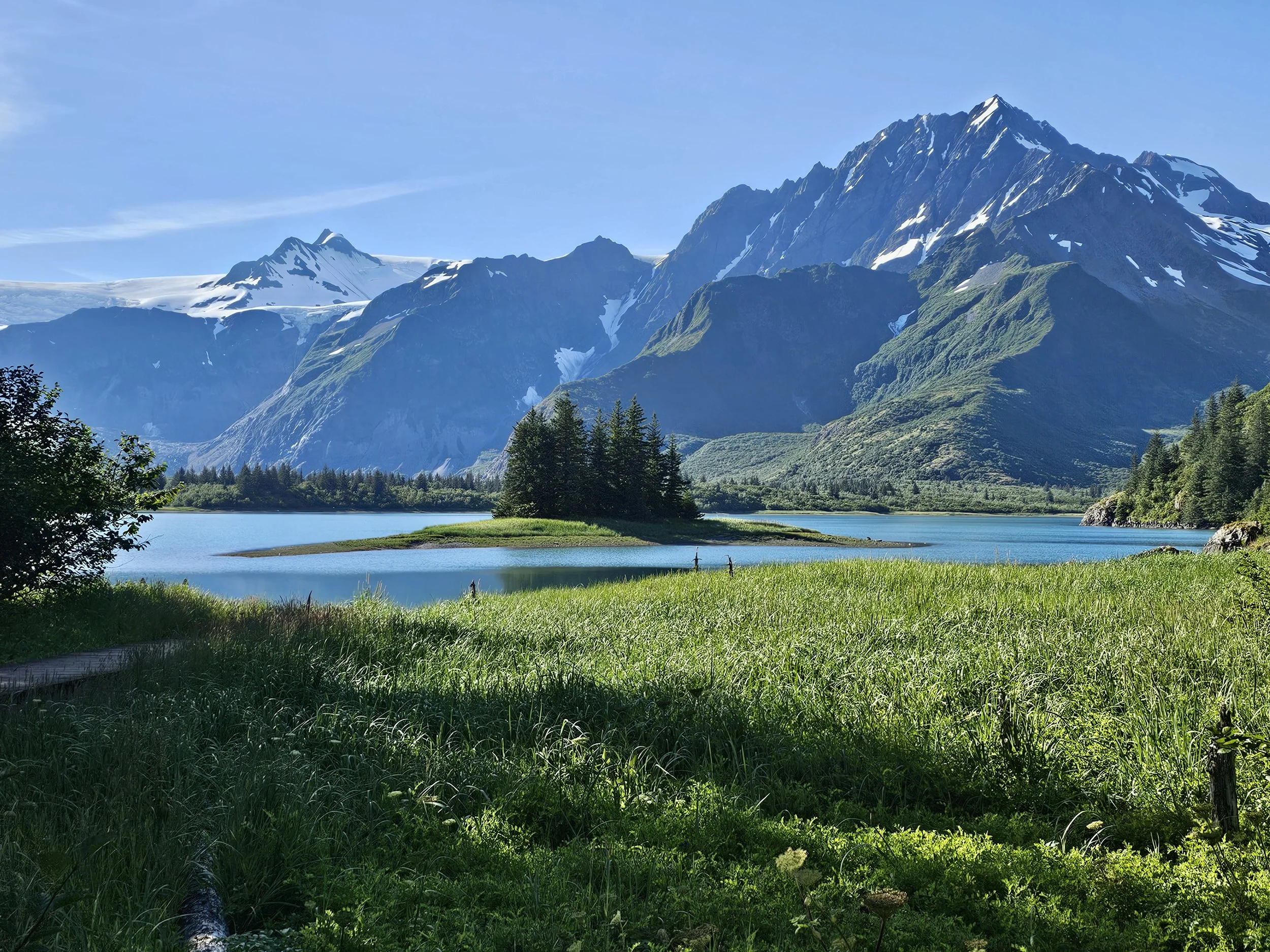Pedersen Lagoon surrounded by green mountains with patches of snow, evergreen trees, and meadow grass under a clear blue sky near Kenai Fjords Glacier Lodge.