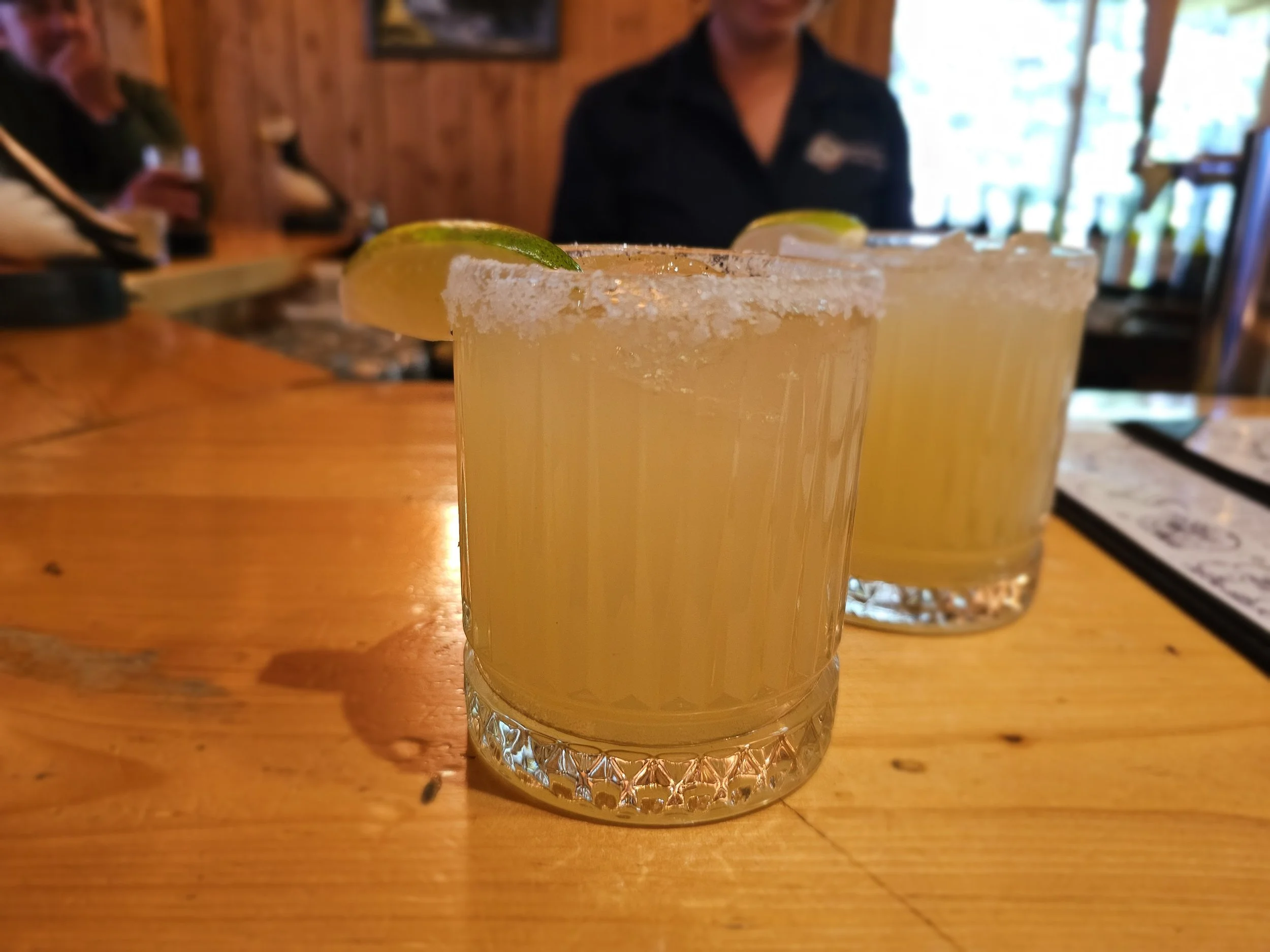 Two margaritas with salted rims and lime wedges on a wooden bar inside Kenai Fjords Glacier Lodge in Alaska.