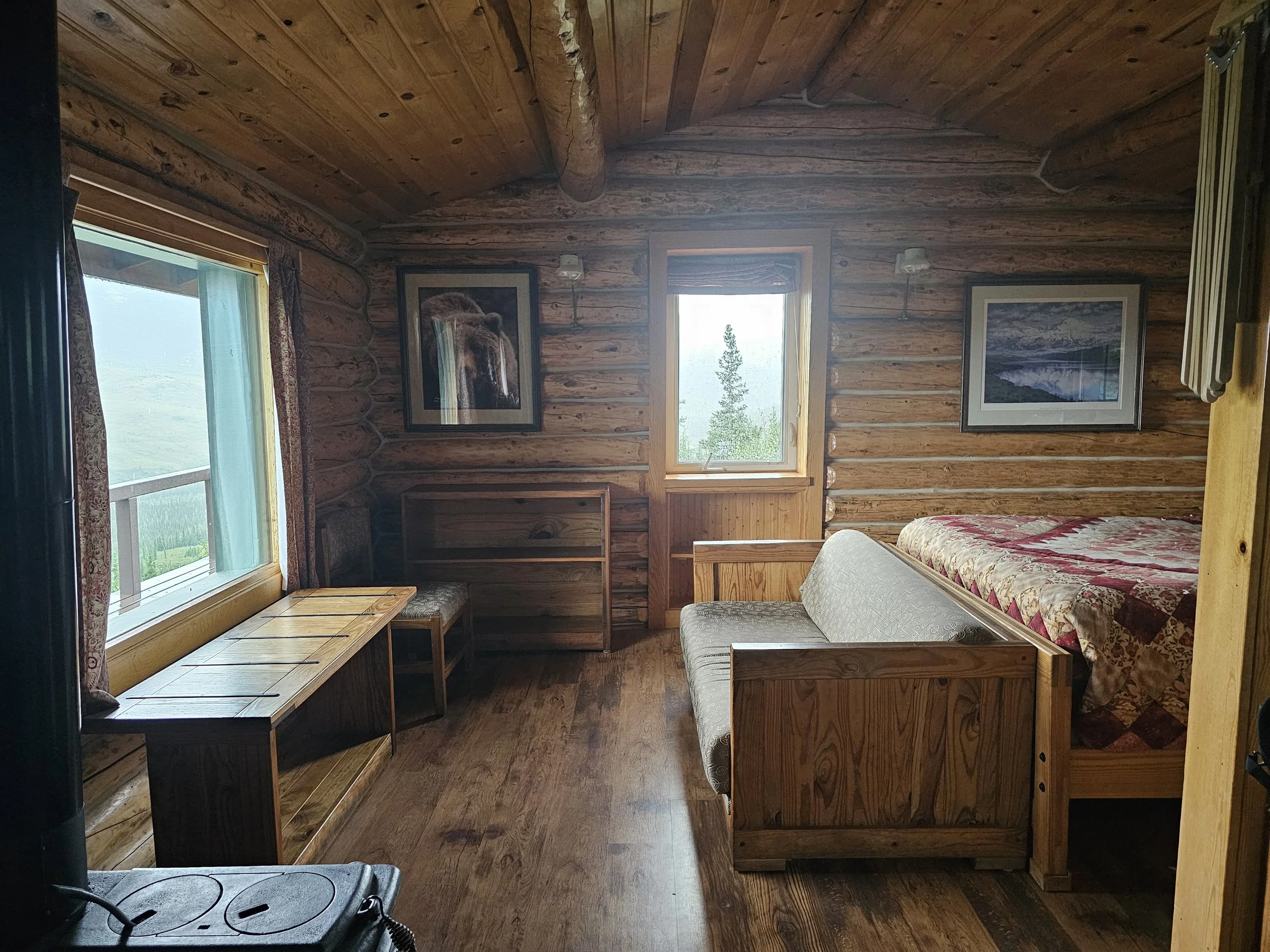 Interior of a Camp Denali cabin with a bed, wooden walls, and windows showing the surrounding landscape.