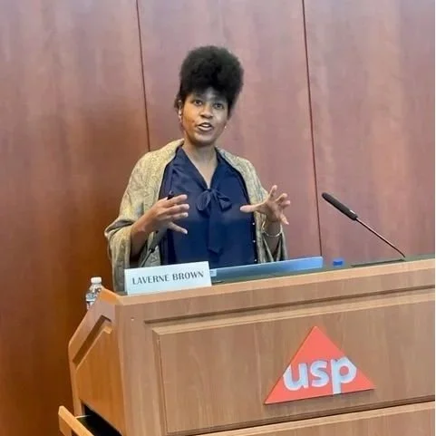 A woman standing at a wooden podium with a sign that says 'LAVERNE BROWN' and a red and white 'USP' logo, speaking into a microphone in a conference room.